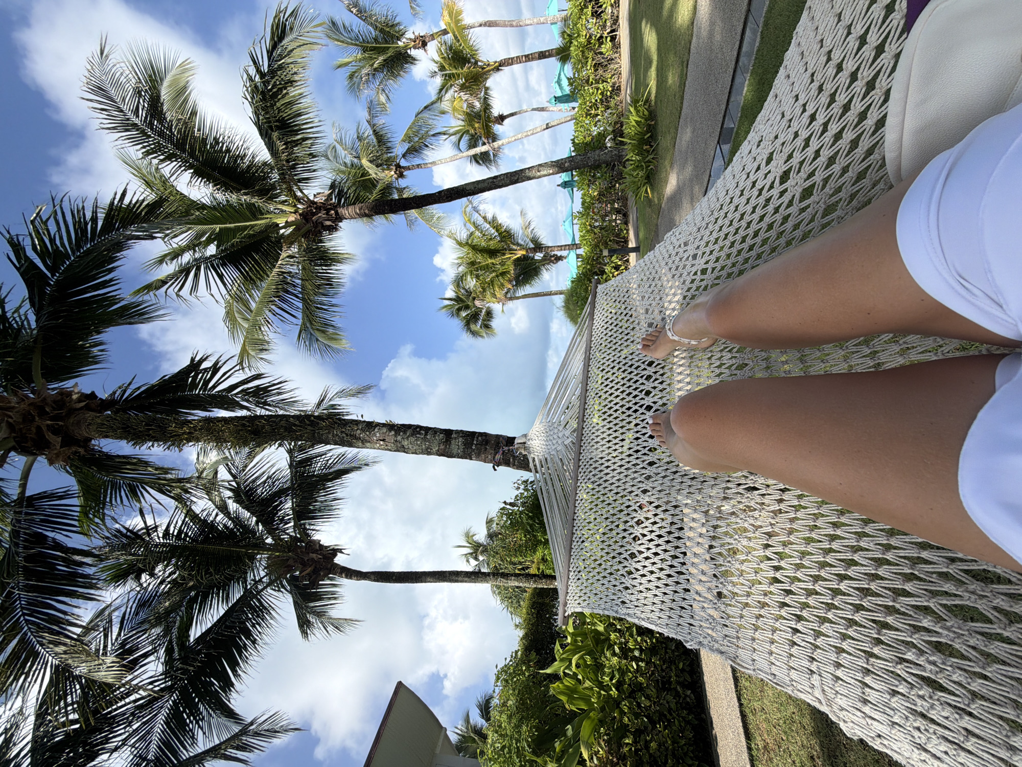 A first-person perspective shot captures a person relaxing in a white macrame hammock, set against a backdrop of lush greenery and palm trees under a bright, partly cloudy sky. The lower half of the person's legs and feet are visible, adorned with white thong sandals. In the distance, turquoise umbrellas and more palm trees suggest a tropical resort or beach location. The scene appears to be outdoors, possibly in a garden area of a resort. The image does not provide specific information to identify the location as Bang Muang, Thailand.