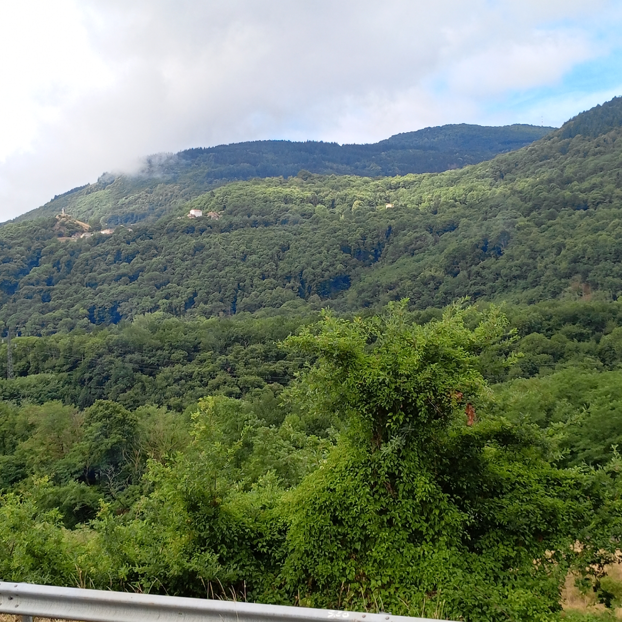 The image depicts a verdant, hilly landscape dominated by dense green trees and foliage. In the distance, a hazy mountain range is visible under a cloudy sky with patches of blue. Several scattered white buildings, likely houses or structures, are nestled within the tree line on the slopes of the mountains. The foreground features a metallic guardrail, suggesting a roadside vantage point. The overall scene conveys a sense of natural, rural beauty, with the lush vegetation indicating a temperate climate. The location is specified as Saint-Paul-de-Jarrat, France, implying a setting within the French countryside, possibly in the Pyrenees foothills given the topography. There are no people or animals visible in the image. The image appears to be a landscape photograph taken during daylight hours.