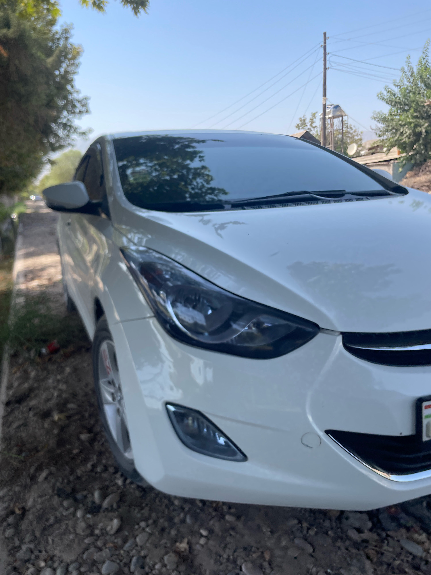 A white Hyundai Elantra is parked on a narrow, unpaved road, possibly in the city of Pasariq, Tajikistan. The car is positioned at an angle, with the front right side closest to the camera. The road is covered in gravel and dirt, with some sparse vegetation growing along the side. In the background, there are trees, a few residential buildings, and power lines. The sky is clear and blue, indicating a sunny day. The image is taken from a low angle, focusing on the car.