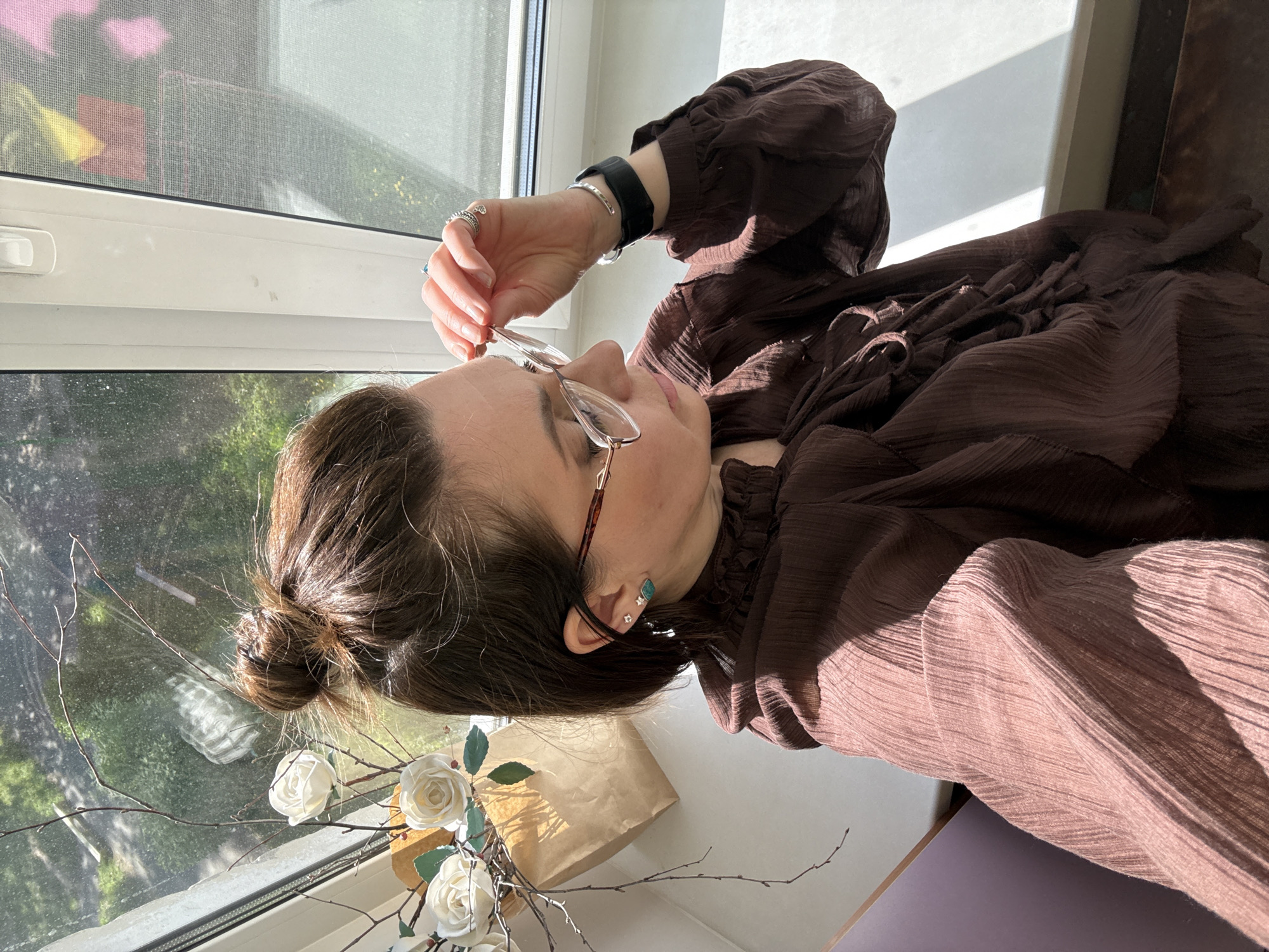 A fair-skinned person with dark hair pulled back in a bun is adjusting their glasses while seated in front of a window. The person is wearing a dark brown, long-sleeved, textured shirt and multiple rings on their fingers. A black smartwatch and a silver bracelet are visible on their left wrist. The background includes a window with sunlight streaming through, a view of trees outside, and a decorative arrangement of artificial white roses and bare branches in a brown paper bag resting on the windowsill. The location is likely in Chaikovsky, Russia.