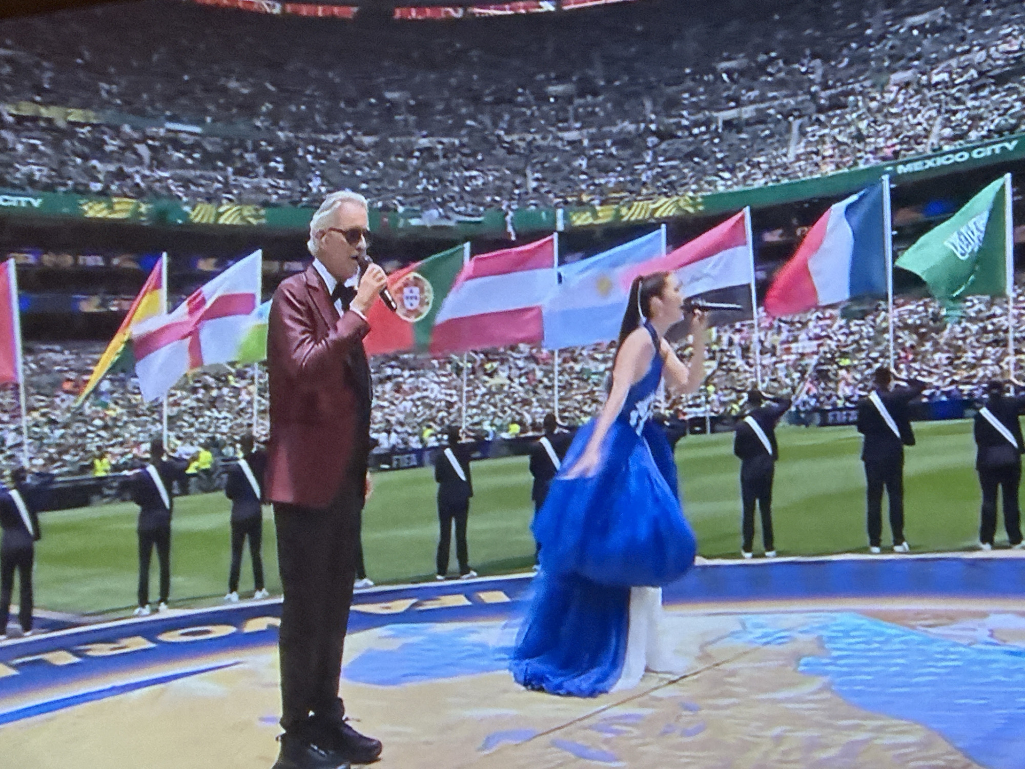 The image depicts an outdoor stadium scene where two singers, a man and a woman, are performing on a large stage area in front of a diverse crowd in the stands. The man, wearing a burgundy suit and sunglasses, is holding a microphone and singing. Beside him, a woman in a flowing blue gown is also singing into a microphone. Several flags are displayed behind the singers, including the flags of Portugal, Spain, Argentina, France, and Saudi Arabia. In the background, a large crowd fills the stadium seating, and a banner reads "MEXICO CITY". Several individuals in dark suits with white sashes stand in formation on the field behind the singers, suggesting a formal event, possibly a sporting ceremony. The ground of the stage is decorated with a large, colorful map-like design.
