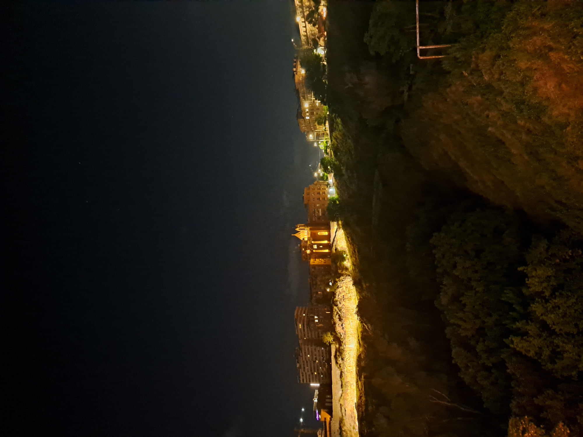 The image depicts a nighttime scene in Yerevan, Armenia. A steep, overgrown hillside slopes downwards from right to left, with sporadic vegetation covering the terrain. Along the top of the hill, illuminated buildings and streetlights stretch out against the night sky. The left side of the image is dominated by a dark, largely featureless sky, with a few faint stars visible. The city's lights create a warm glow against the darkness, highlighting the urban landscape against the natural terrain.