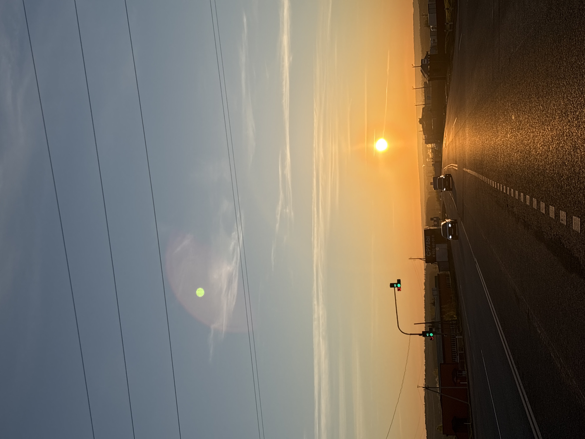 The image depicts a sunrise over a road in Ижевск, Russia. The sky transitions from light blue on the left to orange on the right, where the sun is positioned. Power lines run diagonally across the left side of the frame. A road stretches into the distance, with cars visible. A traffic light with a green light is seen on the side of the road. The road is illuminated by the rising sun, creating a bright reflection. Buildings and trees are visible on the right side of the road.