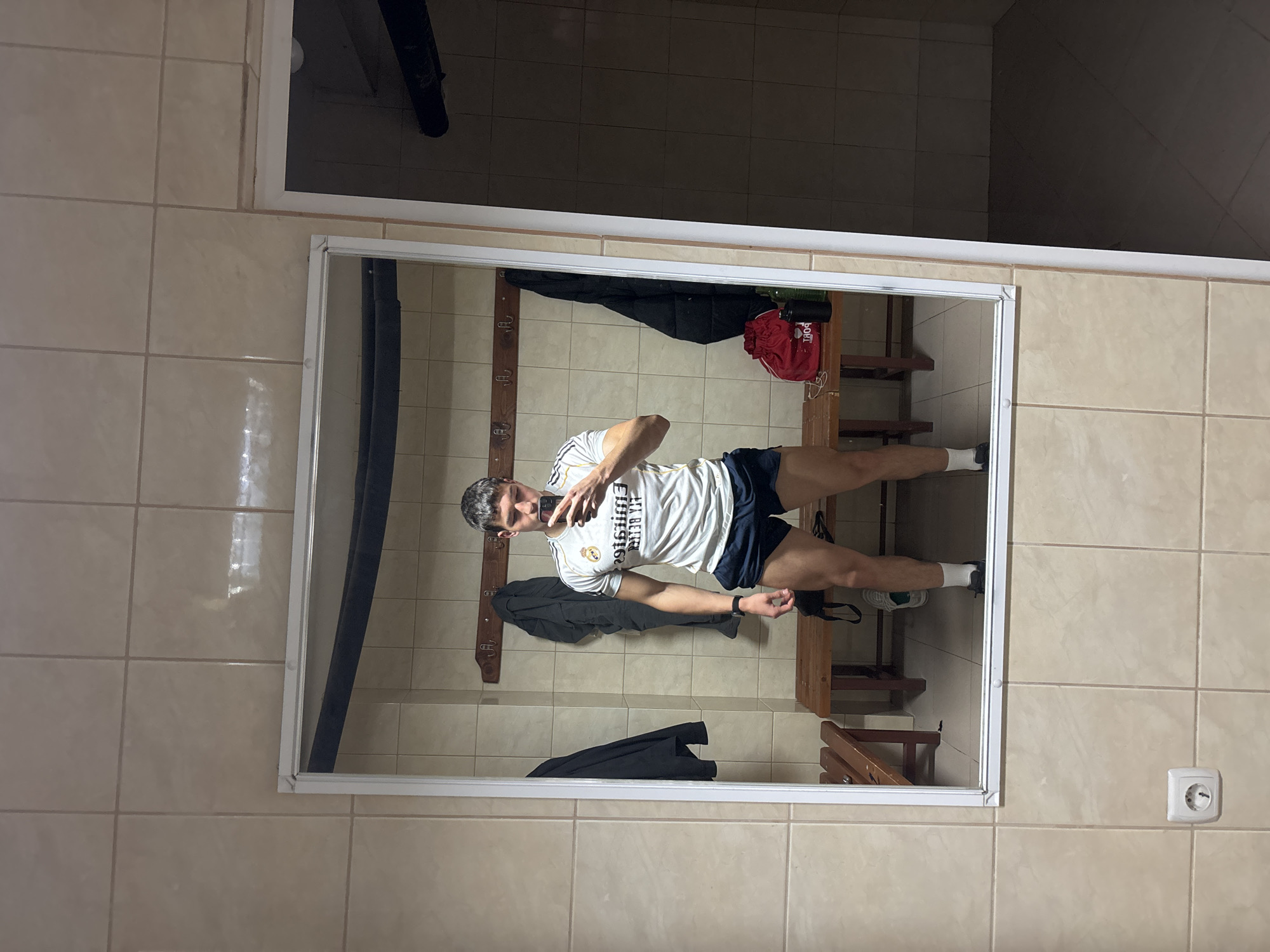 An indoor mirror selfie captures a young man in what appears to be a changing room or locker room in Zlatograd, Bulgaria. The man, with short dark hair, stands facing his reflection, holding a smartphone with both hands to take the picture.
He is dressed in athletic wear, including a white short-sleeved t-shirt with dark stripes on the shoulders, dark shorts, white socks, and sneakers. Visible text on his shirt reads "FLY BETTER" and "EMIRATES," indicating it is likely a sponsored sports jersey, possibly a Real Madrid kit. A dark smartwatch is visible on his left wrist.
The room features light brown or beige tiled walls and floor. Within the mirror's reflection, a wooden panel with several coat hooks is visible behind the man, with a dark jacket hanging on one hook. Further back, wooden benches or shelving units line the wall. A red sports bag with the text "BOFF" is seen hanging in the upper left of the mirror's reflection. Above the mirror's frame and in the unreflected portion of the room, a dark pipe or duct runs along the ceiling. To the right of the mirror, on the tiled wall, there is a white electrical outlet. The mirror itself is framed in white.
The scene suggests an informal moment, likely before or after a sports activity, within a functional and practical setting typical of a sports facility or gym changing area.