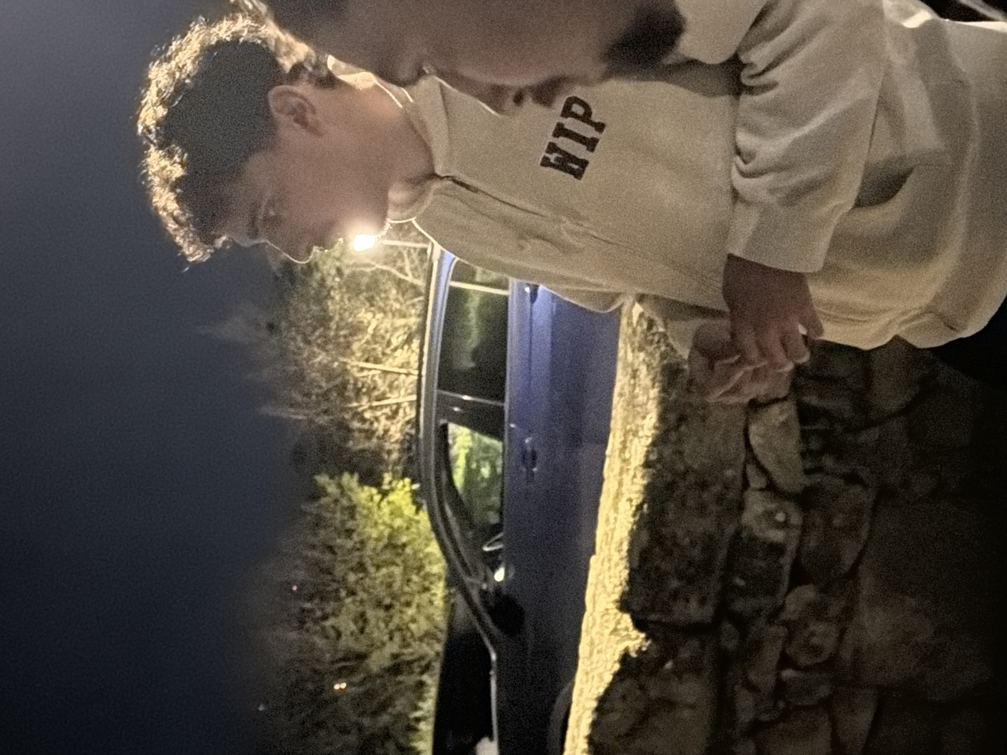 Two individuals are seen in low light conditions, possibly at night, near what appears to be a stone wall. A blue car is parked nearby, and foliage is visible in the background, lit by a streetlight. The location is Vila Real, Portugal. One individual is wearing a light-colored hoodie with the word "HIP" printed on it. The people are standing close to each other, possibly holding hands.