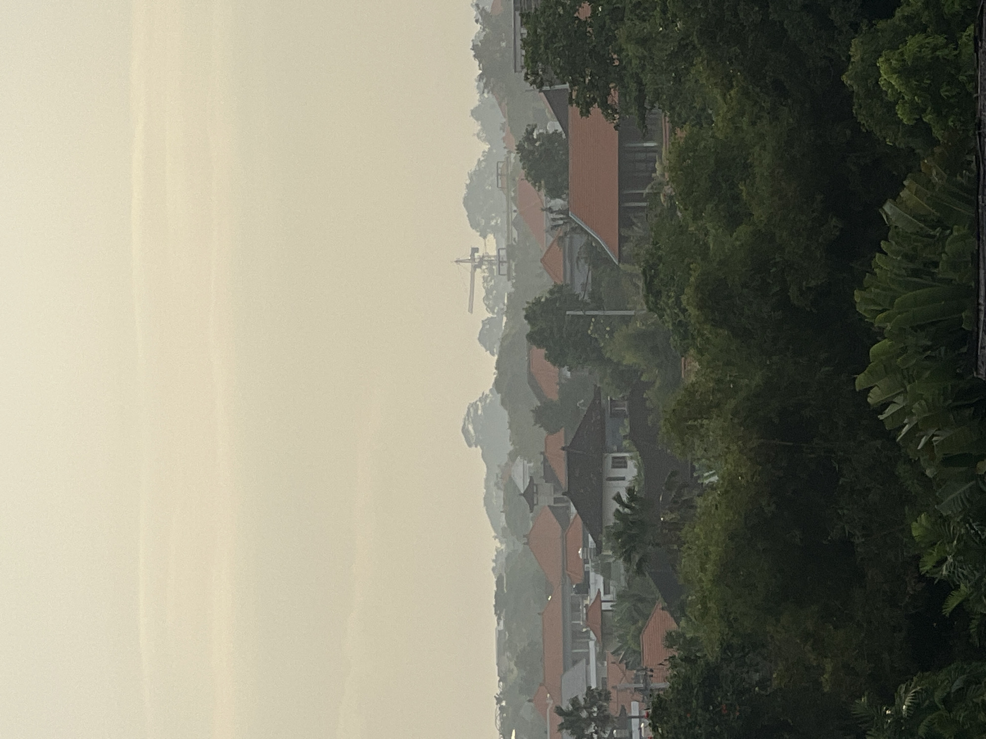 The image depicts a cityscape in Canggu, Indonesia, viewed from an elevated position. The scene is characterized by a dense collection of buildings with orange/red tiled roofs, interspersed with lush green trees and vegetation. The sky is overcast, creating a hazy, muted light across the landscape. In the background, a construction crane is visible, suggesting ongoing development in the area. The overall impression is one of a tropical urban environment partially obscured by weather conditions.