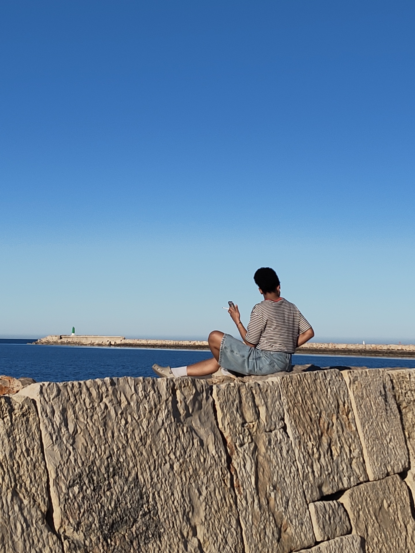 A person sits on a stone wall, their back to the viewer. They are wearing a striped shirt and a blue jean skirt. They are holding a cigarette in their right hand. The sea is behind the wall with a breakwater, a green lighthouse, and a cloud visible. The sky is clear and blue. The location is most likely in Dénia, Spain, based on the breakwater and lighthouse style.