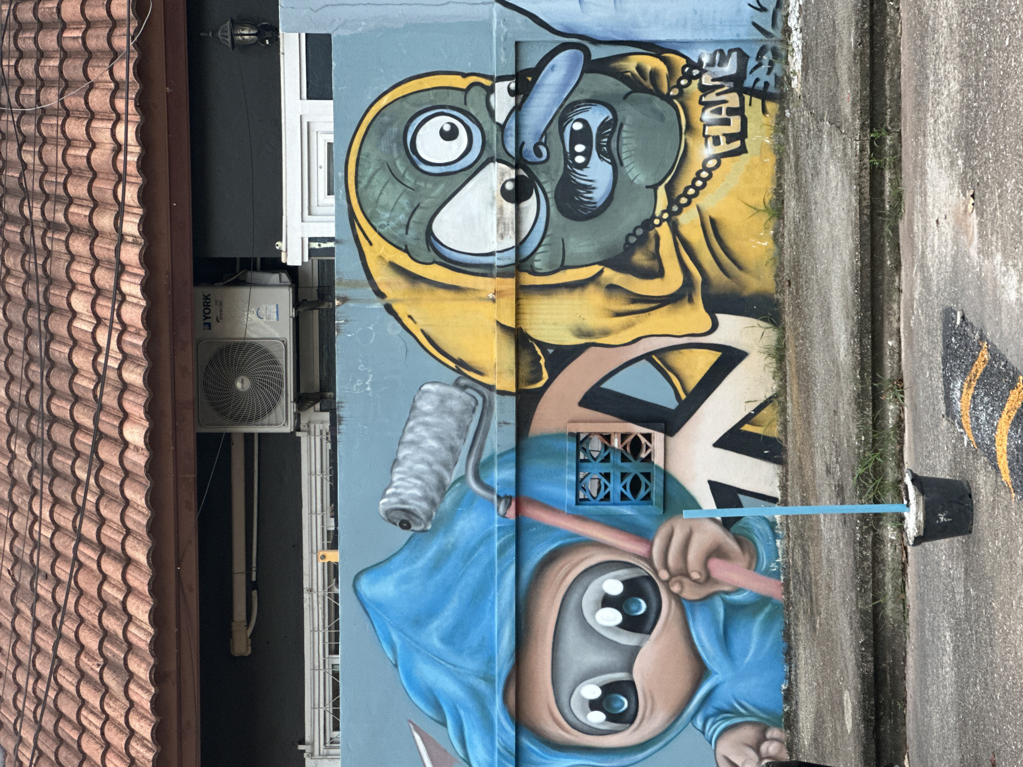 This media file captures a vibrant mural on a light blue wall in Kathu, Thailand. The mural features two stylized cartoon characters, each with large, expressive eyes. The upper character wears a yellow hooded jacket and has a green face with a blue nose and tongue. A beaded chain is visible around its neck, and the word "FLAME" is spray-painted in bold, blocky letters above and to its right. The lower character is depicted wearing a blue hooded garment and holding a pink rod connected to a paint roller. Its face is predominantly brown with large, wide eyes that have white irises and blue pupils.
The setting appears to be an urban or semi-urban environment, possibly an alleyway or the side of a building. To the left of the mural, a portion of a brown tiled roof is visible, suggesting a residential or commercial building. An outdoor air conditioning unit is mounted on the wall below the roofline. The mural is painted over a textured blue wall, with parts of the wall appearing weathered or uneven. To the right of the mural, a concrete surface transitions into an asphalt road, with some patches of grass growing in the cracks and along the edges. A black bucket with some white material spilling out and a blue stick resting on it is positioned on the concrete. The overall impression is one of urban art brightening a somewhat utilitarian space. The activity depicted is the act of painting, represented by the paint roller held by the lower character. No specific time of day or weather conditions are explicitly indicated, but the lighting suggests it is daytime.