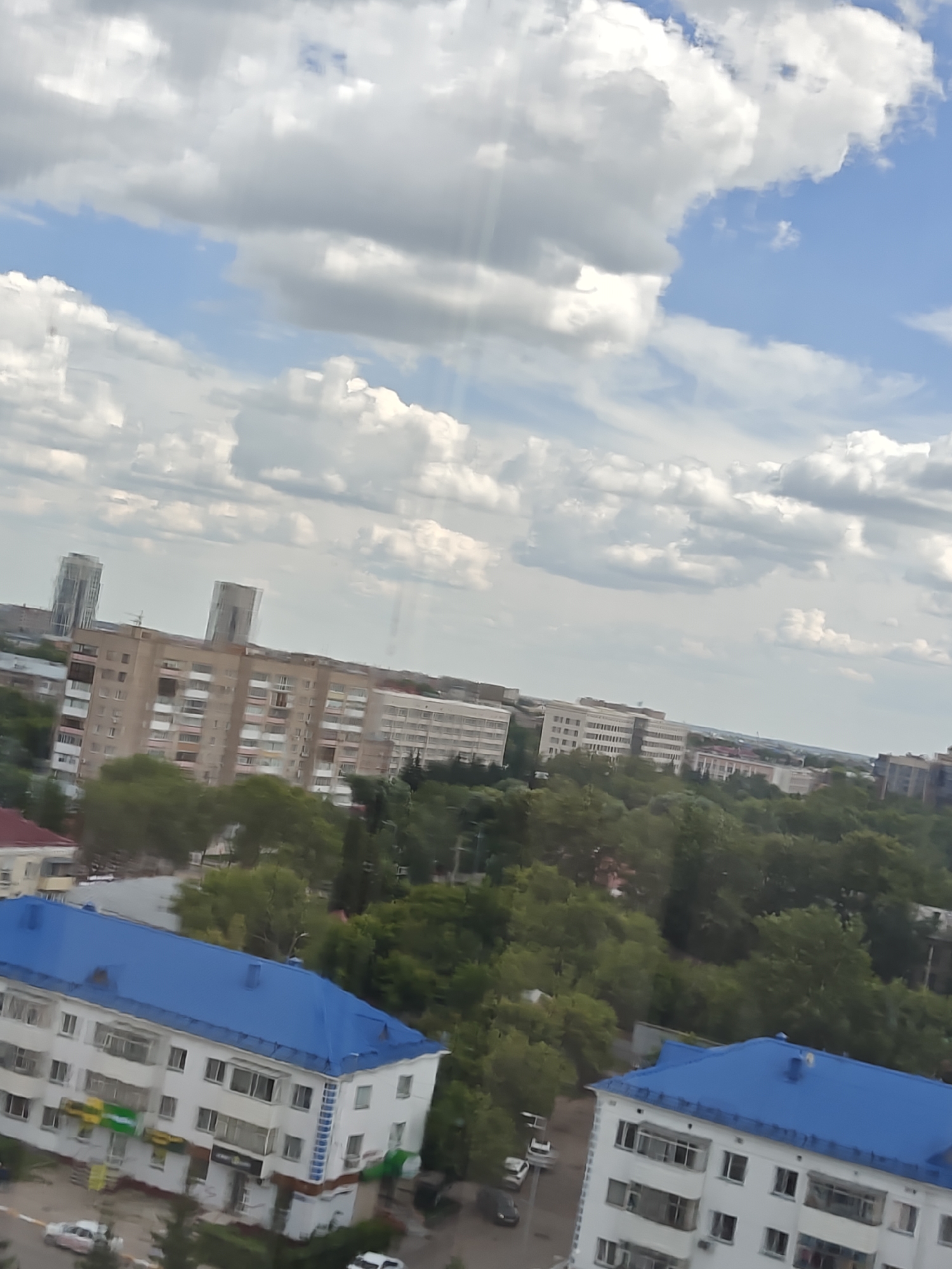 The image shows a high-angle view of a city skyline, with a prominent blue-roofed apartment building in the foreground. The sky is partly cloudy with white cumulus clouds and patches of blue sky. In the background, there are several other apartment buildings of varying heights and architectural styles, some with balconies and visible windows. A line of trees separates the foreground and background buildings, creating a green buffer. The city appears to be Petropavl, Kazakhstan, based on the distinctive architecture of the buildings, particularly the taller, modern structures in the distance. The lighting suggests it is daytime.