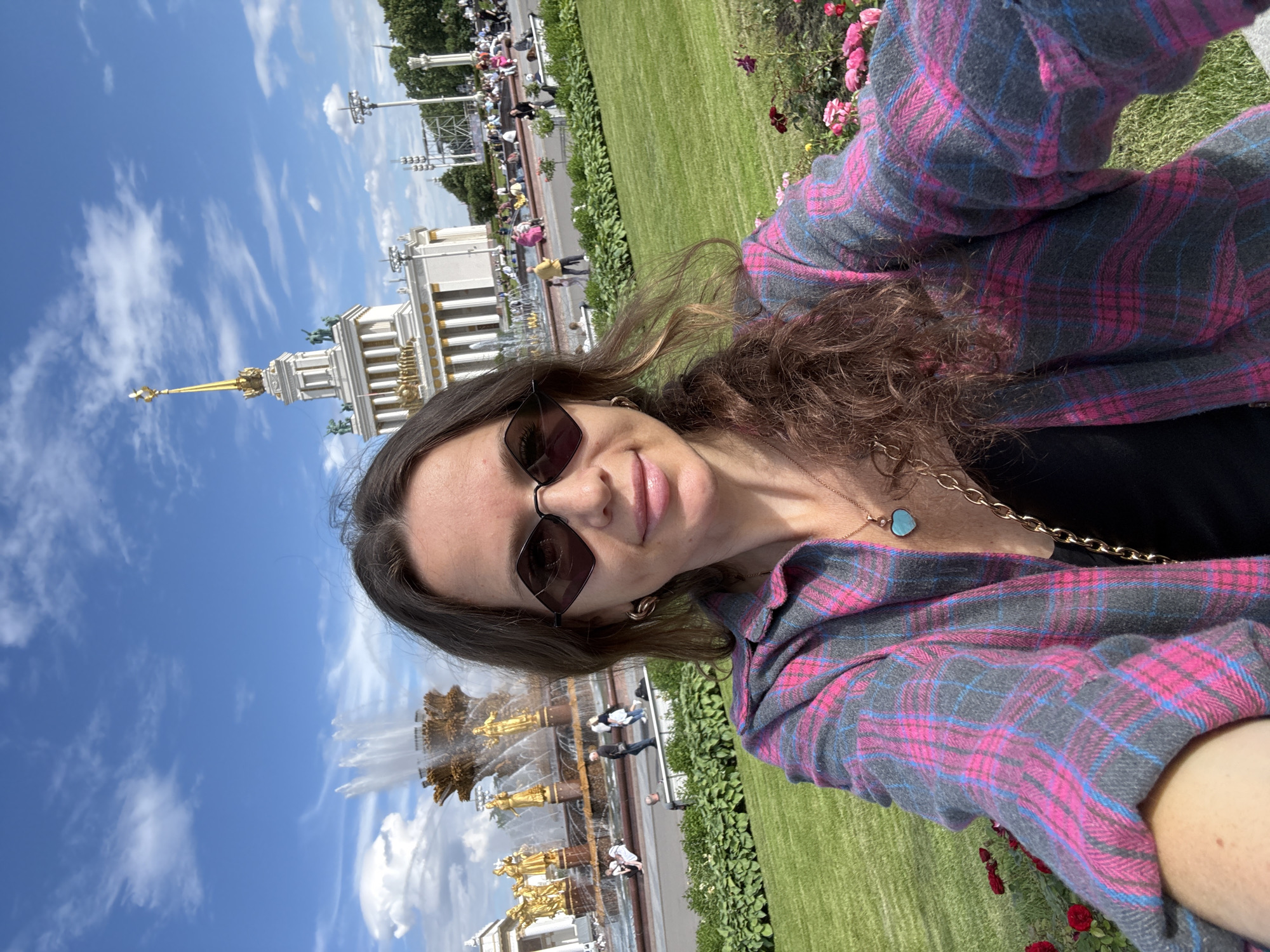 The image is a selfie taken by a woman in a park. The woman is wearing sunglasses and a pink and gray plaid shirt. She has brown hair and is smiling. In the background, there is a large, ornate fountain with golden statues. Behind the fountain, there is a white building with a golden spire, and beyond that, a green lawn and trees. The sky is blue with scattered clouds. The time of day appears to be daytime, and the weather is sunny. The location is likely Moscow, Russia, given the architecture and style of the fountain. The overall mood is cheerful and relaxed, suggesting a tourist enjoying a day out in a scenic location.