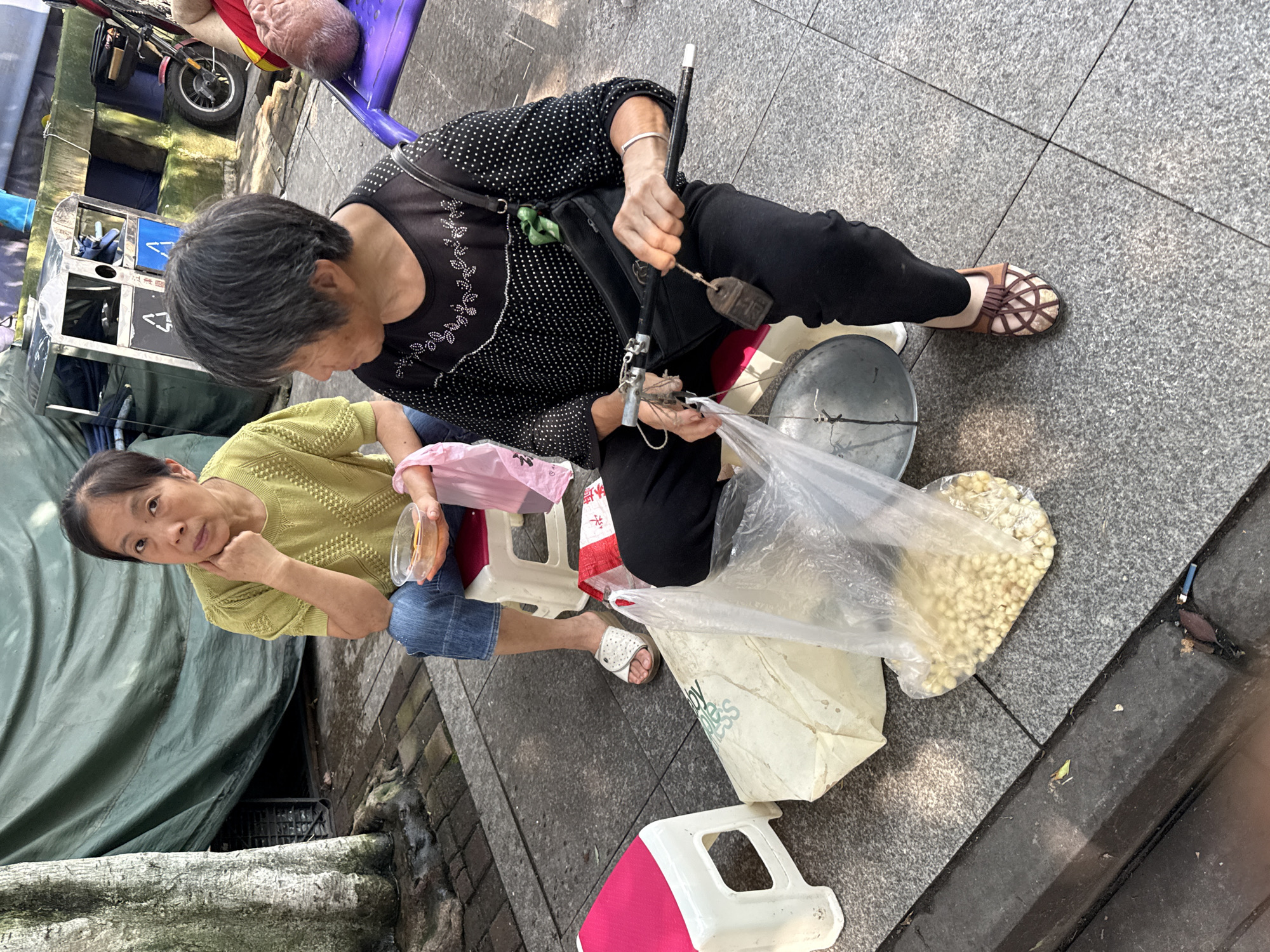 Two elderly Asian women are seated on small stools on a paved sidewalk in Jiangjin, China. The woman on the left, wearing a lime green knitted shirt and blue jeans, holds a clear plastic bowl with food. The woman on the right, in a black polka-dot shirt and black pants, is holding a weighing scale with a handle. Beside her is a plastic bag filled with small, light-colored spherical objects, likely food items being weighed for sale. In the background, a large green tarp covers some structures, and various objects, including a bicycle and a trash bin, are visible. The scene appears to be a street-side vending operation.