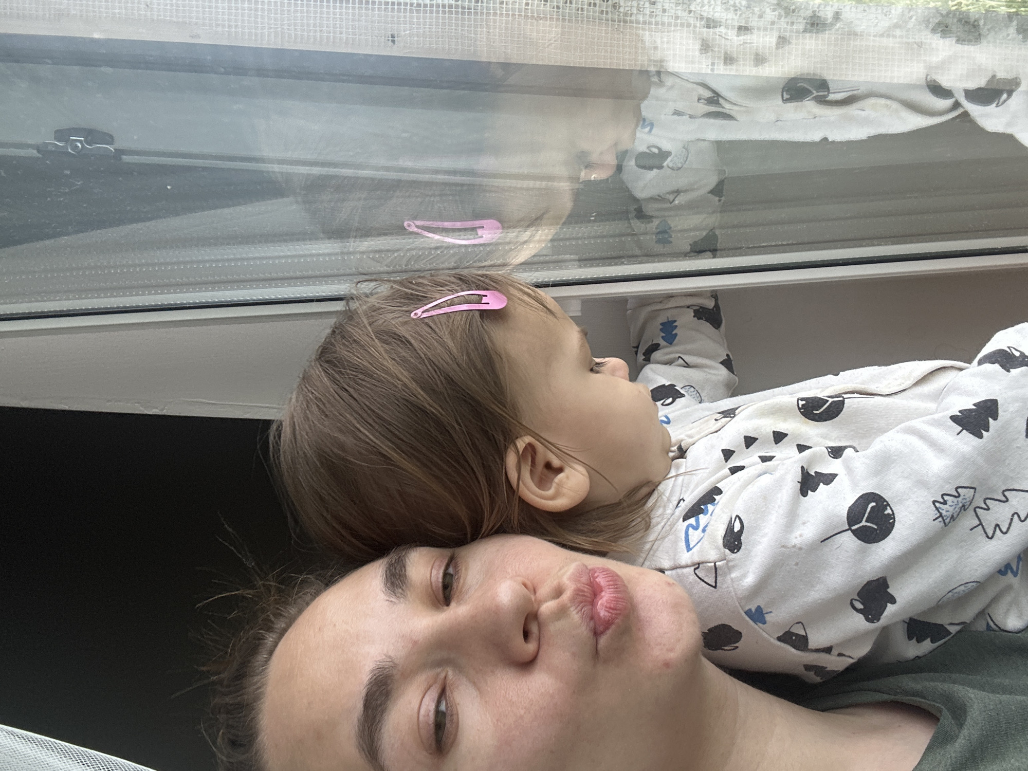 A woman and a child are close together, with the child lying on the woman. The woman is facing the camera and has dark hair pulled back, with her face angled slightly upwards. She is making a pouting expression. The child has brown hair with a pink hair clip. The child is wearing a white pajama top with a tree and cloud pattern. Above them is a window with a reflection of the child and their clothing. There's a window latch visible. Behind and beside the people, there is darkness. It is not possible to identify the location of the image.