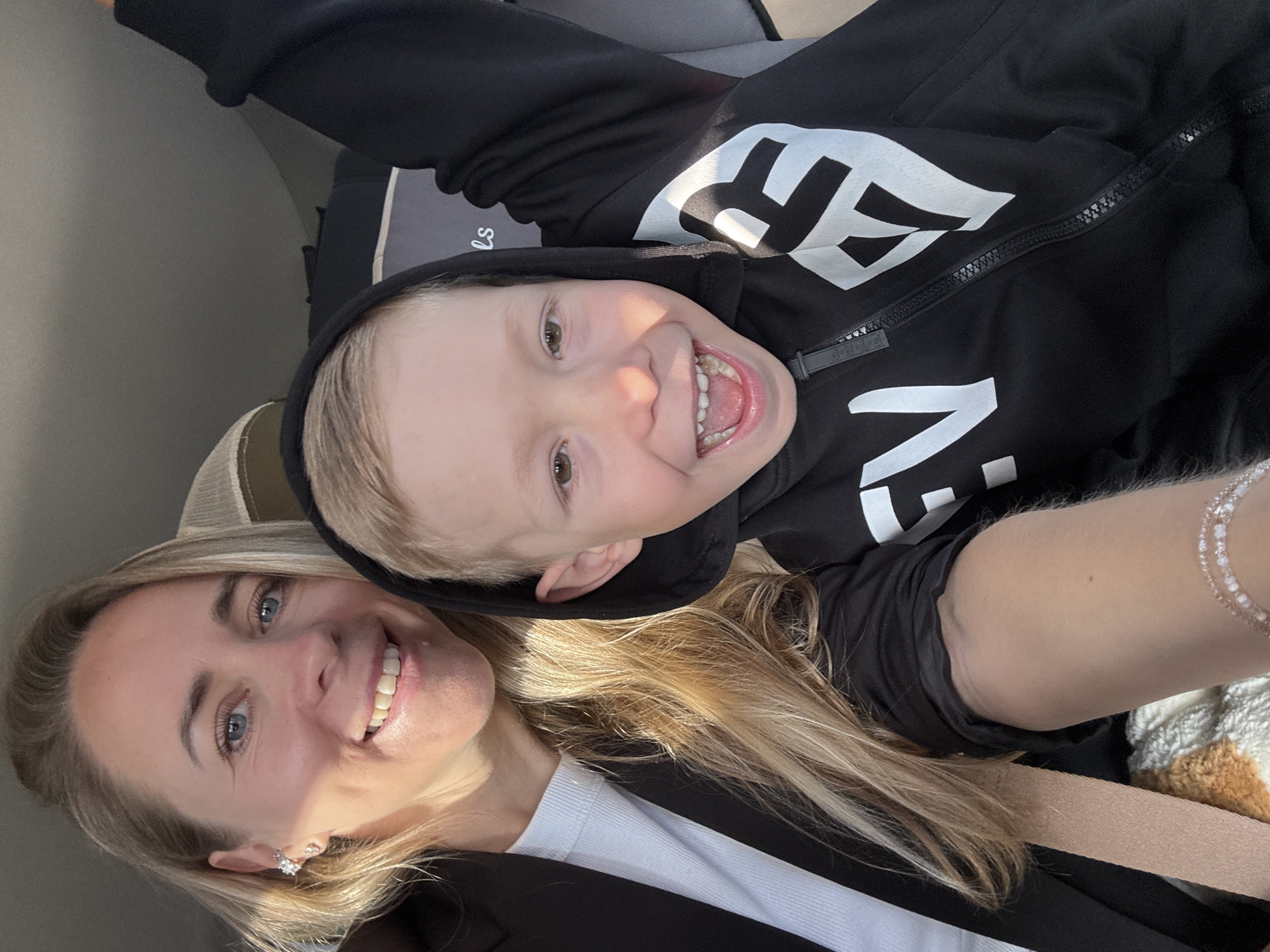 In this selfie, a blonde woman and a young boy are in a car. The woman is on the left, smiling and wearing a black blazer over a white top. She has blue eyes and a diamond earring. The boy is on the right, with blond hair and a wide smile, wearing a black hooded sweatshirt with white lettering. His right arm is raised. The background suggests they are in the back seat of a car, with a car seat visible. The lighting indicates it is daytime. The image does not provide any information about the city of Chaikovsky, Russia.