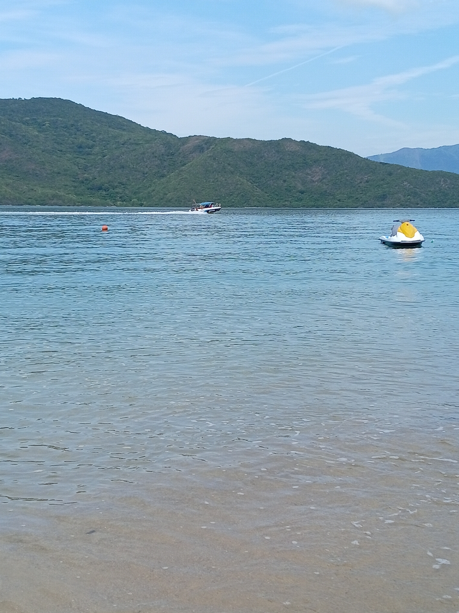 The image depicts a serene coastal scene at Hoa Thang, Vietnam. The foreground features the shallow, sandy bottom of the ocean, with gentle waves lapping onto the shore. In the middle ground, the clear blue water stretches out, reflecting the sky. Two vessels are visible on the water: a jet ski, primarily white with a yellow seat, is stationary in the right side of the frame, while a speed boat, with occupants visible, is moving rapidly towards the left in the upper middle of the frame, leaving a wake. A small orange buoy is also present in the water, between the shore and the speed boat. In the background, a lush, green, and mountainous landscape dominates the horizon, meeting a clear blue sky with faint white clouds. The overall atmosphere is tranquil and scenic.