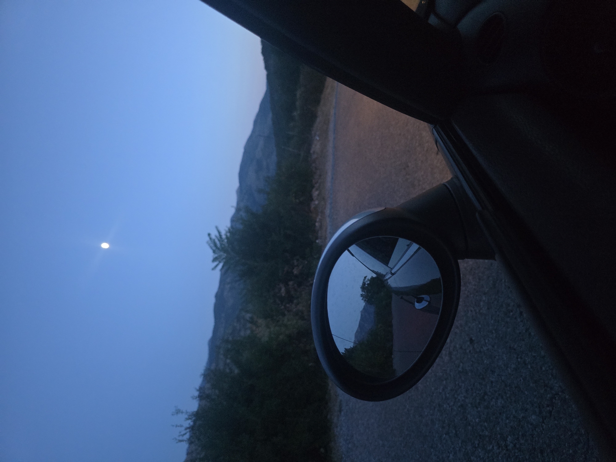 The image shows a rearview mirror of a car, reflecting a twilight landscape with a crescent moon in the sky. The car appears to be parked or slowly moving on a gravel road, with dark green trees and rolling hills in the background. The scene is likely in Klopot, Montenegro, given the mountainous terrain and sparse vegetation typical of the region. The overall mood is serene and tranquil, evoking a sense of peaceful solitude.