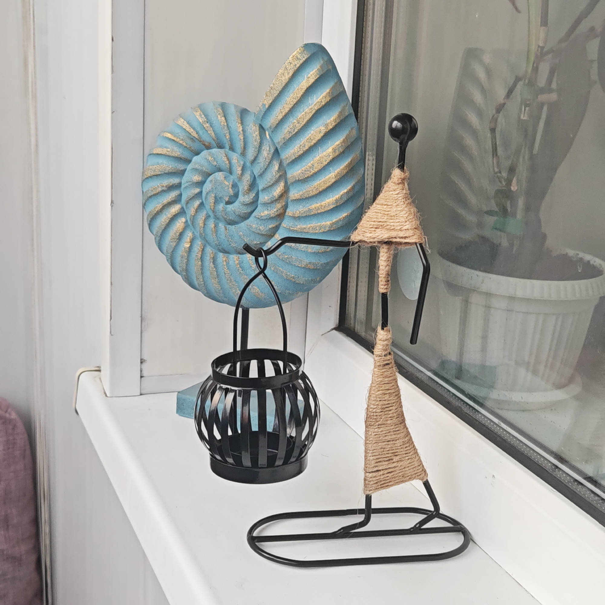 On a white windowsill, likely indoors, a light blue and gold nautilus shell sculpture stands alongside a black metal lantern and a metal sculpture of a person wrapped in brown twine. The window behind the objects reflects a potted plant. There is no indication of location within Ketoshi, Russia. No people are visible, and no actions or interactions are occurring.