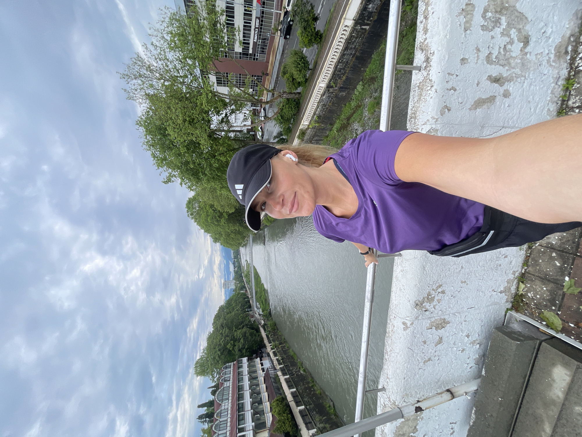 A woman is seen in a selfie, leaning against the railing of a bridge overlooking a river. She is wearing a purple t-shirt, black shorts, and a black Adidas baseball cap, along with white wireless earbuds.
The bridge appears to be aging with peeling white paint. Below the bridge, a river flows with buildings and trees lining its banks. There are buildings and trees visible in the background as well.
The setting is Sochi, Russia, based on the architectural style of the buildings and the urban riverfront landscape. The sky is cloudy.