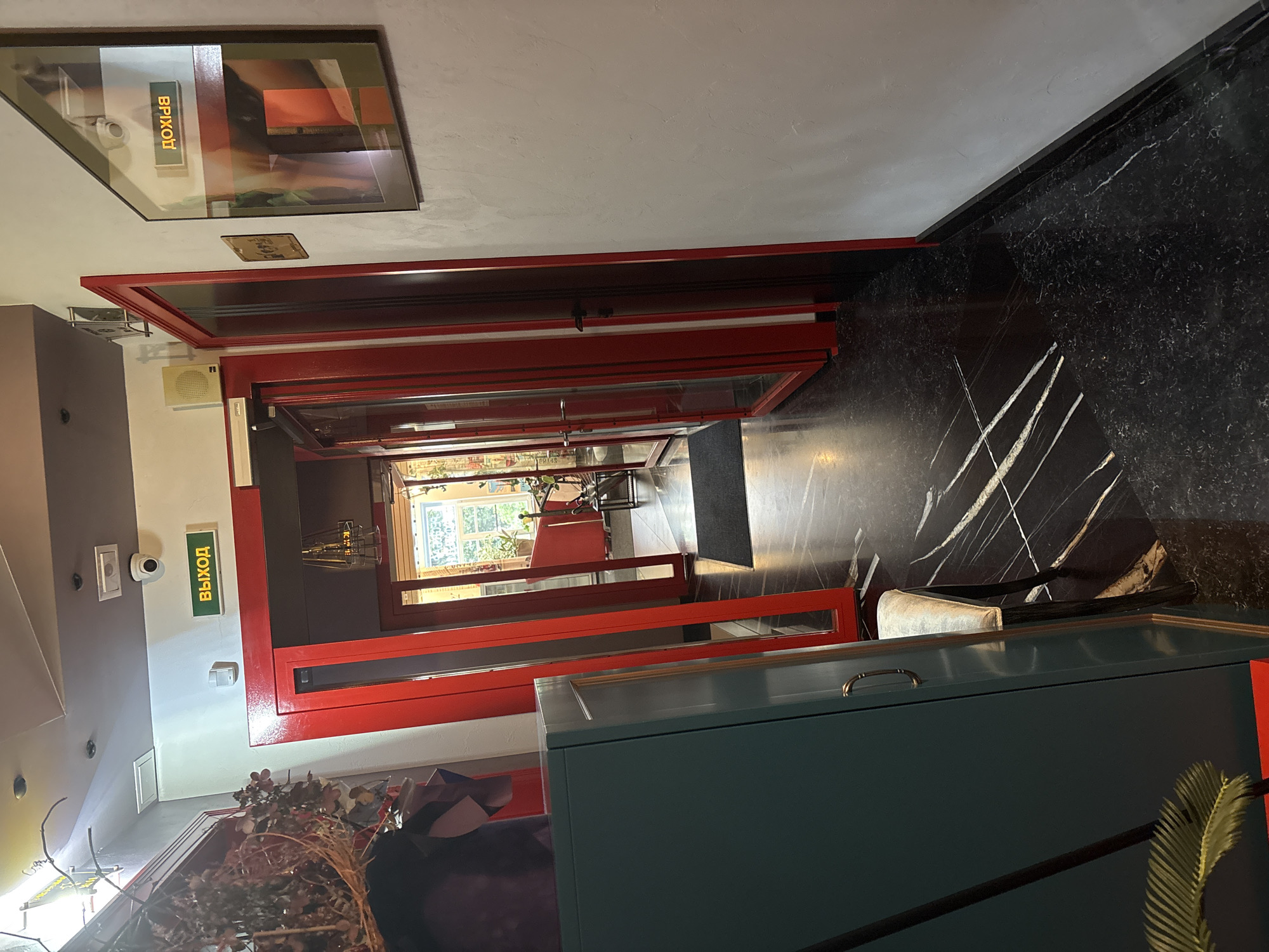 This is a photograph taken inside what appears to be a modern establishment, possibly a restaurant or cafe, in Saint Petersburg, Russia. The image is oriented vertically, with the camera looking down a hallway.
The hallway features a striking black marble floor with white veins, which contrasts with the bright red frame of the double doors that line one side. These doors have glass panels, offering glimpses into other rooms. On the left wall, there is a white surface with a security camera and a rectangular vent. Further down the hallway, a green illuminated sign with the word "ВЫХОД" (meaning "EXIT" in Russian) is prominently displayed. Above this sign, a framed piece of art hangs on the wall.
The scene suggests an indoor setting, likely during daytime, as natural light is visible from the window at the end of the hallway, which overlooks greenery. There are no people visible in the frame. The overall impression is one of a clean, contemporary space with bold design choices. The red elements, the marble floor, and the artwork contribute to a sophisticated yet potentially lively atmosphere. The presence of the "EXIT" sign indicates it's a public space with designated exits.