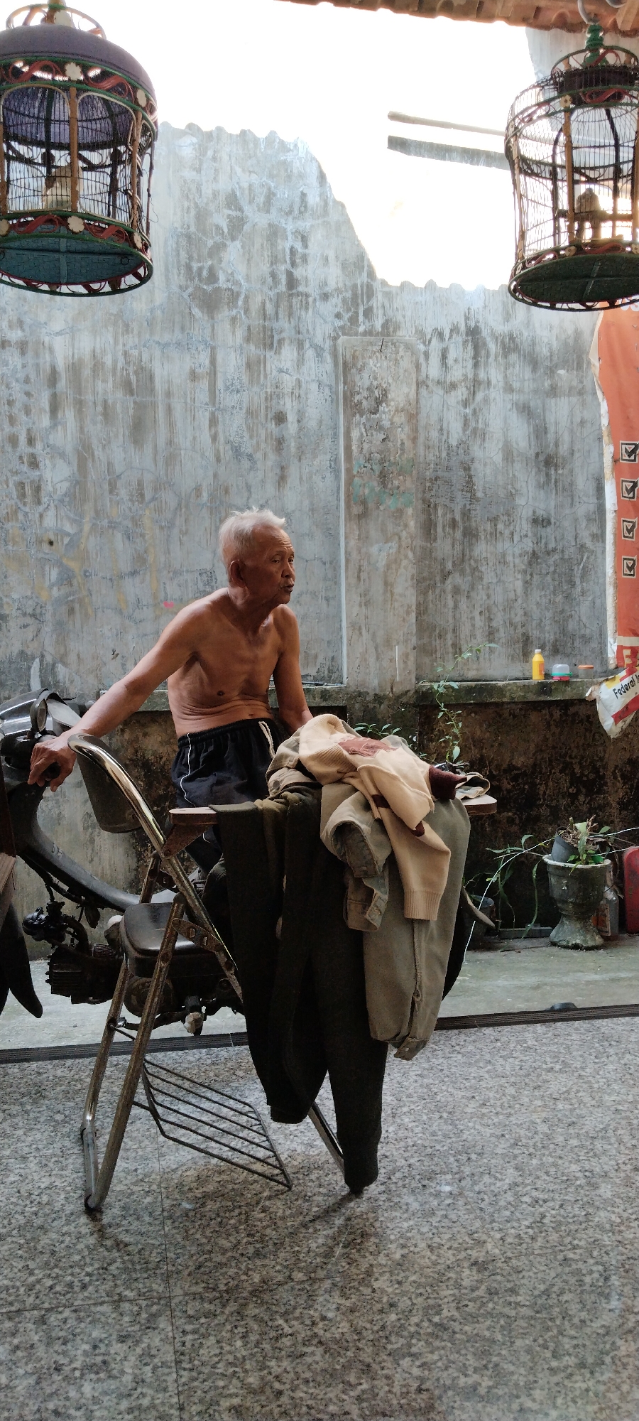 A shirtless, elderly Asian man with white hair sits on a metal chair next to a motorcycle. He is wearing black shorts with white trim. The chair is positioned in front of a weathered, textured concrete wall. On the seat of the chair, several articles of clothing are draped: a light-colored sweater with a brown pattern, a dark green jacket, and a pair of dark pants. To the left of the man, a part of the motorcycle's handlebars and front section is visible. Above and to the sides of the scene, decorative bird cages hang from unseen supports. The ground is covered in a speckled, grey and white stone-like material. In the background, potted plants and a few small objects are visible on a ledge against the wall. A bright, white light source, likely the sun, illuminates the scene from the upper right.