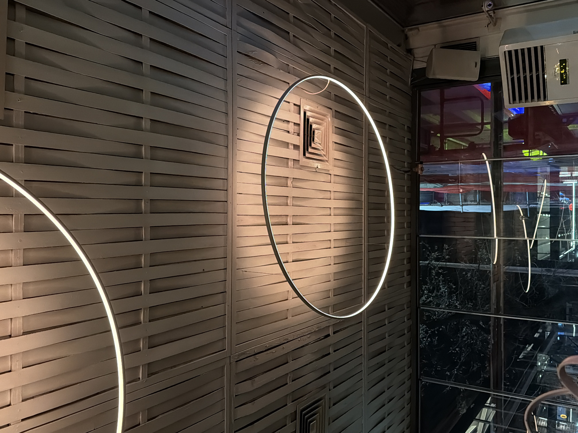 The image displays the interior ceiling and upper wall section of Mr. B. Takeaway & Pizza, located at bul. "Knyaz Boris I" 11, 9000 Varna Center, Varna, Bulgaria. The ceiling is composed of light-toned, possibly grey or beige, narrow rectangular strips arranged in an interlocking grid pattern. Two circular LED light fixtures are suspended from this ceiling; one complete fixture is prominent in the lower-middle of the frame, emitting a bright white light, and a partial second fixture is visible in the upper-left corner. A square, multi-layered air vent is centrally positioned within the lower complete circular light fixture. Another rectangular air vent is partially visible in the upper-left. The lower portion of the image features large windows, reflecting the dark nighttime exterior which shows indistinct tree branches and various colored artificial lights. On the right side, adjacent to the window, a white wall-mounted speaker is affixed. Below the speaker, a floor-standing white air conditioning unit with horizontal grilles and a digital display showing "16.0" is present. A small, white security camera is mounted on the wall above and to the right of the air conditioning unit. A curved, light-colored object, possibly a furniture armrest, is partially visible in the bottom-left corner. No individuals are depicted in the scene.