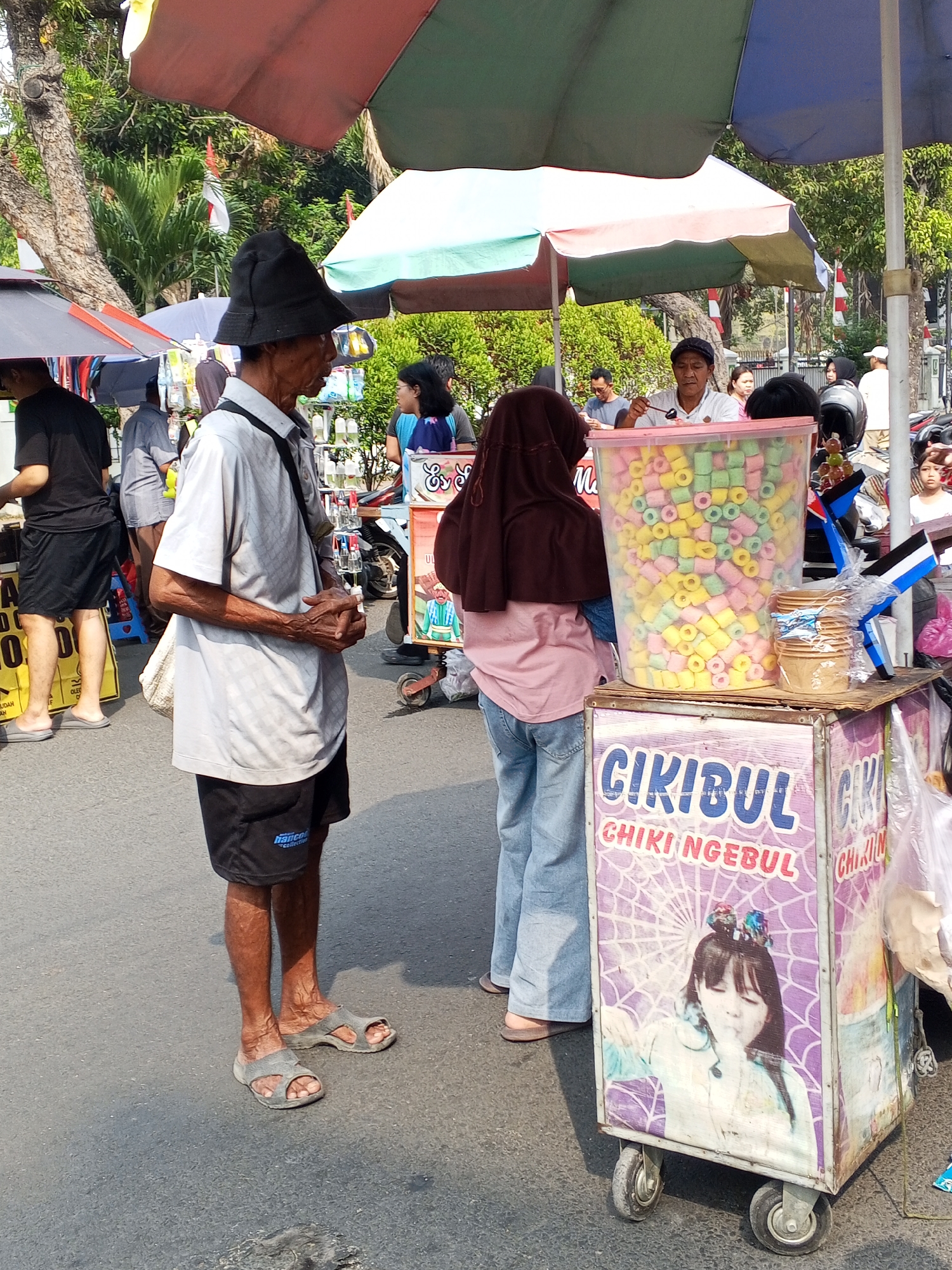 The image depicts a street scene in Serang, Indonesia, likely near a market or busy thoroughfare. In the foreground, an elderly man wearing a black hat, a light-colored collared shirt, and black shorts stands facing right, his hands clasped in front of him. He is positioned next to a colorful vendor cart filled with brightly colored snacks, possibly puffed corn snacks, in a large transparent plastic container. The cart is decorated with images and text, including "CIKIBUL CHIKI NGEBUL" and a picture of a young girl. A woman wearing a hijab and a pink shirt, along with light-blue jeans, stands to the right of the cart, appearing to interact with the vendor. Behind them, other people and vendor stalls are visible under colorful umbrellas, suggesting a commercial area. The ground is paved, and the overall atmosphere appears to be that of a typical bustling street market in Indonesia.