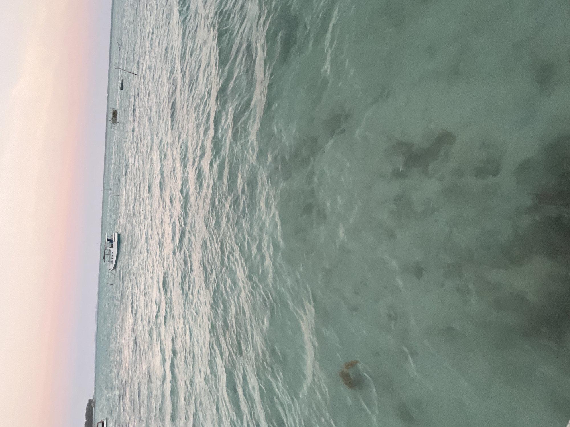 The image shows a serene seascape at dusk in Legonipah, Indonesia. The sky displays a gradient of soft pink and orange hues, transitioning to a pale blue horizon. The calm sea is a tranquil turquoise, with gentle ripples disturbing its surface. In the distance, a small white boat with a canopy is anchored, its silhouette sharp against the pastel sky. Further out, another rudimentary structure, possibly a fishing raft or marker, is visible. The foreground of the water is exceptionally clear, revealing the sandy seabed below, dotted with scattered coral fragments and a solitary piece of debris, appearing orange and circular, floating just beneath the surface. The overall atmosphere is peaceful and tranquil, capturing the quiet beauty of the coastal environment as day gives way to evening.