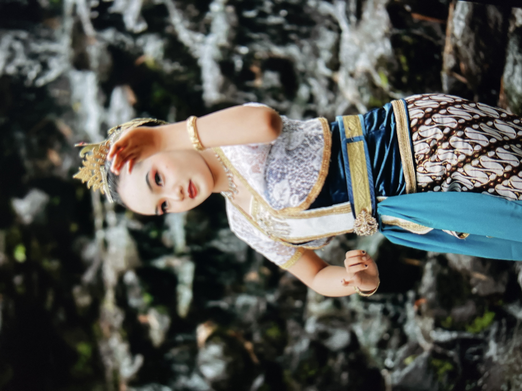 A young woman, adorned in traditional Javanese attire, performs a dance against a backdrop of a rocky waterfall. She wears a gold crown, a white lace top with gold trim, a dark blue sash, and a patterned batik sarong. Her makeup is bold, with red lipstick and winged eyeliner. Her pose is graceful, with one hand raised to her head and the other extended forward. The waterfall, likely located near Cirahayu, Indonesia, provides a natural, textured environment. The lighting is diffused, suggesting an overcast day or shaded area.