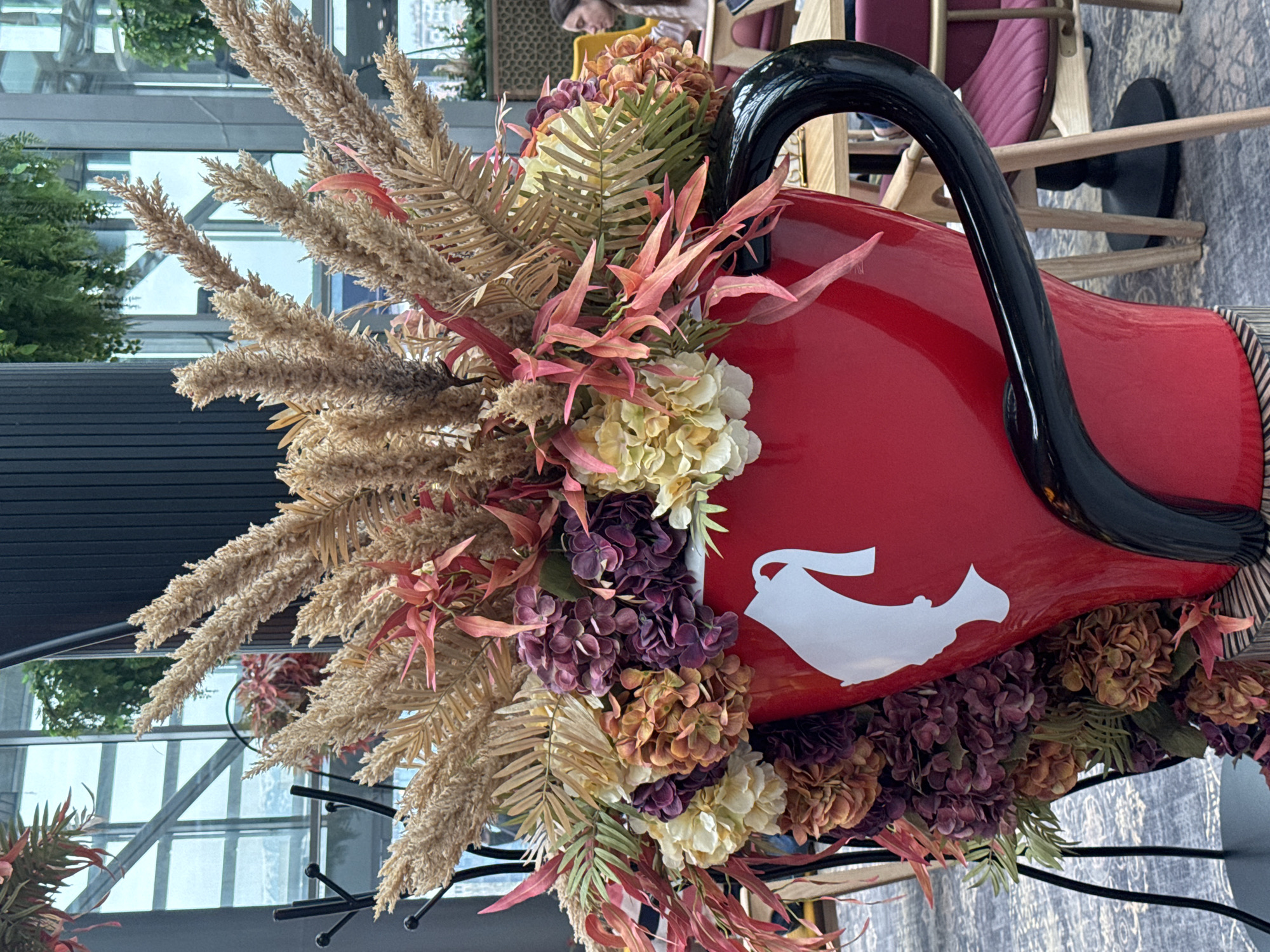 The image shows a large, red, oversized teacup as a decorative element, filled with various dried flowers and greenery. The teacup features a white silhouette of a teapot on its side and has a large, black handle. The arrangement includes beige, brown, and reddish-brown dried plants with what appears to be hydrangeas. The teacup is in a space with large windows, reflecting other parts of the interior. A person can be seen in the background at a table with chairs, implying a restaurant or cafe setting. Given the presence of Bulgarian text in the image details, this scene could potentially be located in the city of Sofia, Bulgaria.