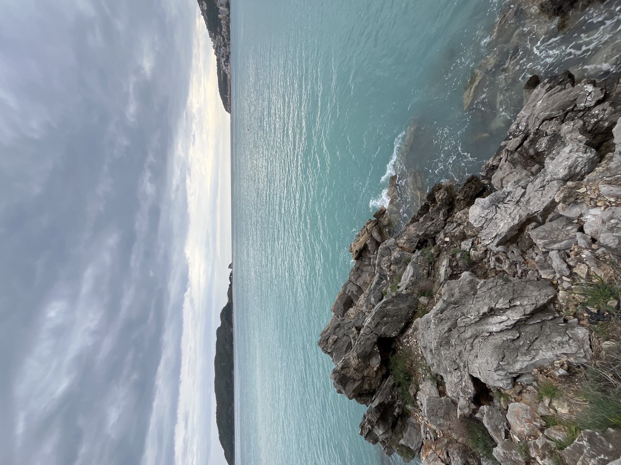 A landscape scene shows a rocky shoreline abutting the turquoise sea under a cloudy sky. The rocky area is in the lower right of the frame, with craggy grey rocks and some sparse vegetation. The sea stretches horizontally across the middle of the image, exhibiting gentle ripples on its surface. A strip of land is visible in the far distance on the left side of the image, with buildings suggestive of a town or city, likely the city of Budva, Montenegro. The overcast sky is filled with grey clouds in varying patterns. The image orientation is rotated ninety degrees, with the sky on the left and the rock on the right.