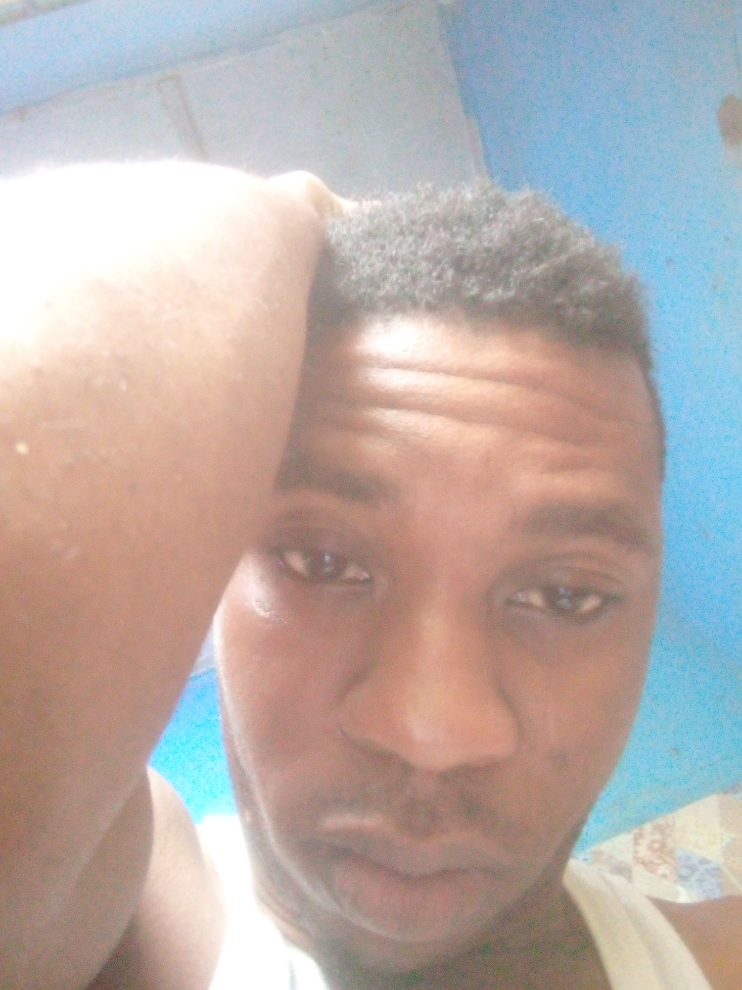 A close-up selfie of a dark-skinned man with short, dark hair. He is wearing a white sleeveless shirt. His left hand is on his head. The background is a blue wall. The location is possibly a residential area in Ikorodu, Nigeria, but there is no visual confirmation.