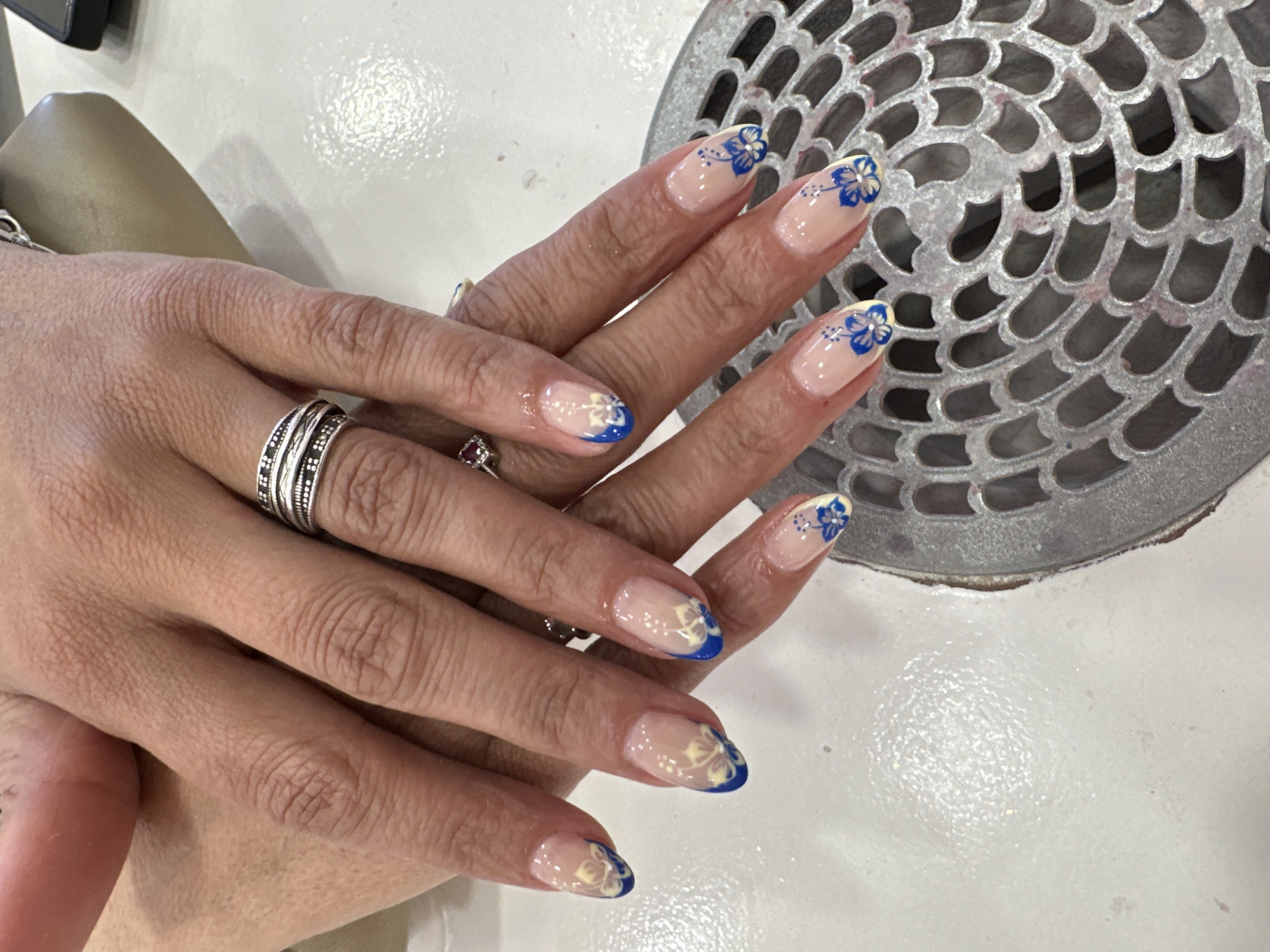 The image shows a close-up of two hands with manicured nails. The nails have a clear base with a blue tip and a floral design. The hands are adorned with rings. One ring is a wide silver band with intricate patterns, and the other appears to be a smaller ring with a red stone. The background includes a silver-colored, perforated metal object and a light-colored surface. No location information or specifics about Sofia, Bulgaria are present in the image.