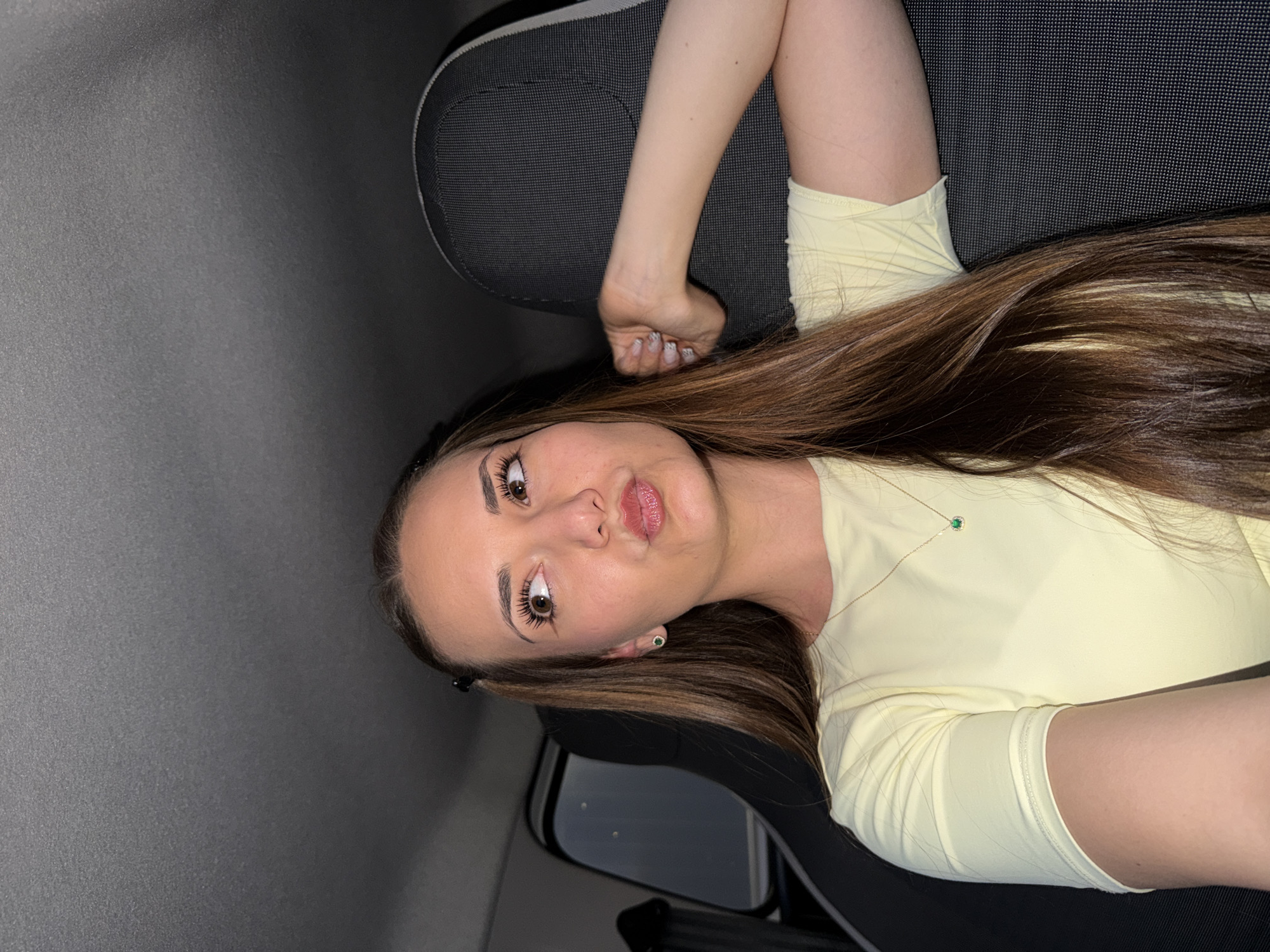 A young woman with long, brown hair is sitting in a car. She is wearing a pale yellow shirt and a delicate necklace with a small green pendant. She has makeup on, including eyeliner and lip gloss. The car's interior is dark, with a gray headliner and dark seats. The image does not provide any information regarding the location in Sofia, Bulgaria.