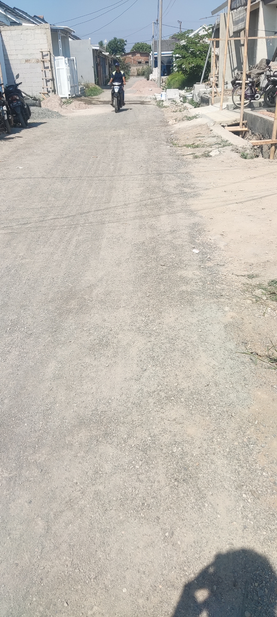 A person rides a motorcycle on a dirt road in Gebang, Indonesia. The road is lined with construction materials and unfinished buildings. The sky is clear and blue.