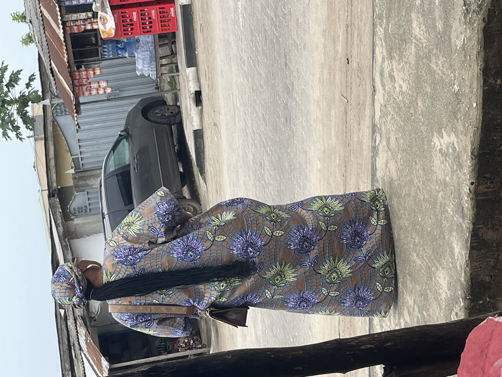 In Badagry, Nigeria, a woman wearing a patterned dress and headwrap stands on a concrete surface near a building with a corrugated metal roof. A gray car is partially visible next to the building, which appears to be a shop with goods displayed. Crates and bottles are stacked in the background.