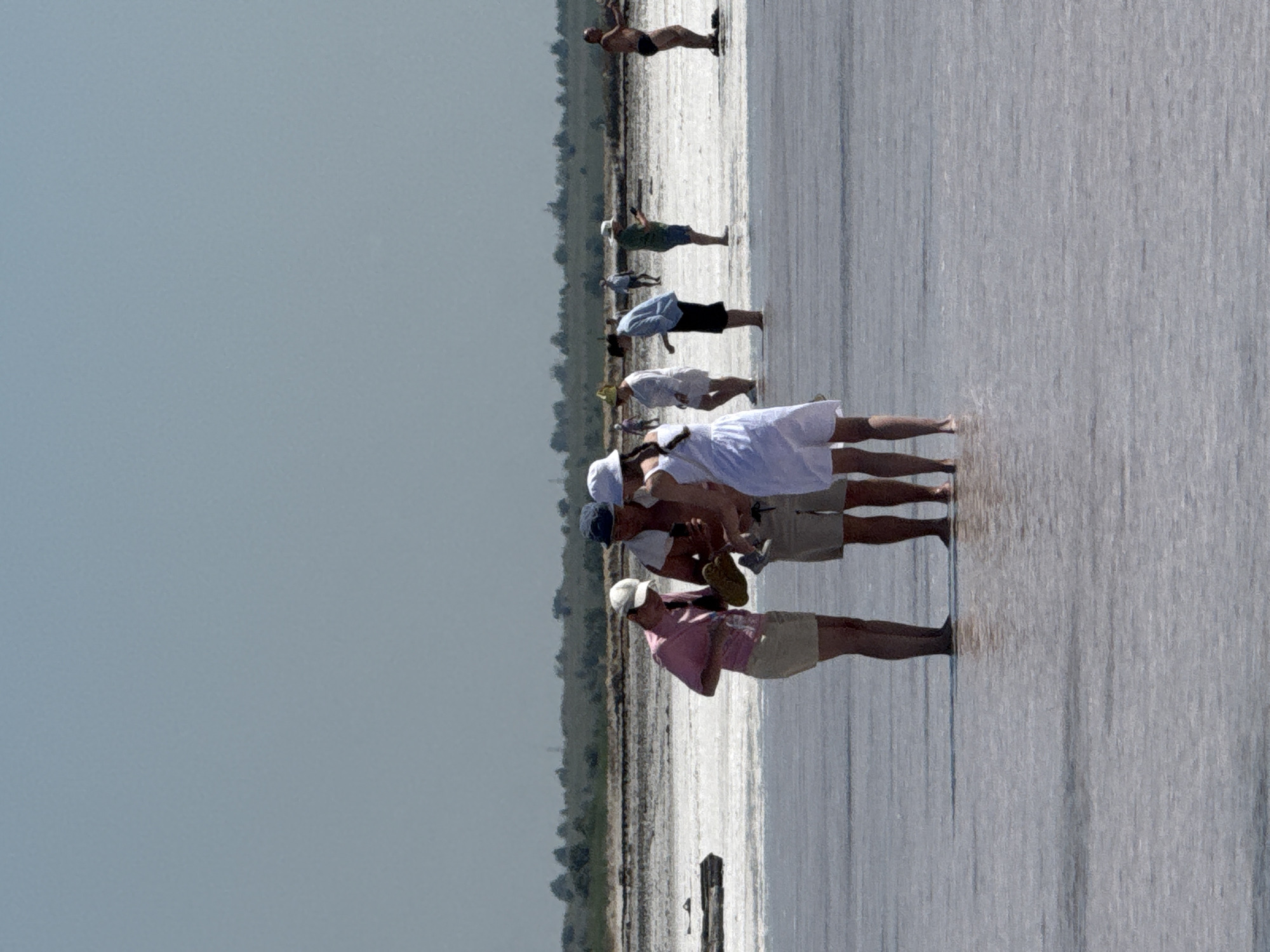 In Bursol', Russia, a group of people are wading in the shallow waters of a salt lake under a hazy sky. In the foreground, three individuals stand together. A woman in a pink shirt and beige shorts holds a pair of brown sandals. Beside her, a man in a blue cap and tan shorts looks at his phone, holding a sandal in his other hand. To his right, a woman in a white hat and white dress is partially obscured. Further in the background, other individuals are scattered across the salt flats, some walking, some observing the water. The landscape is characterized by flat, white salt deposits and a distant line of green vegetation.