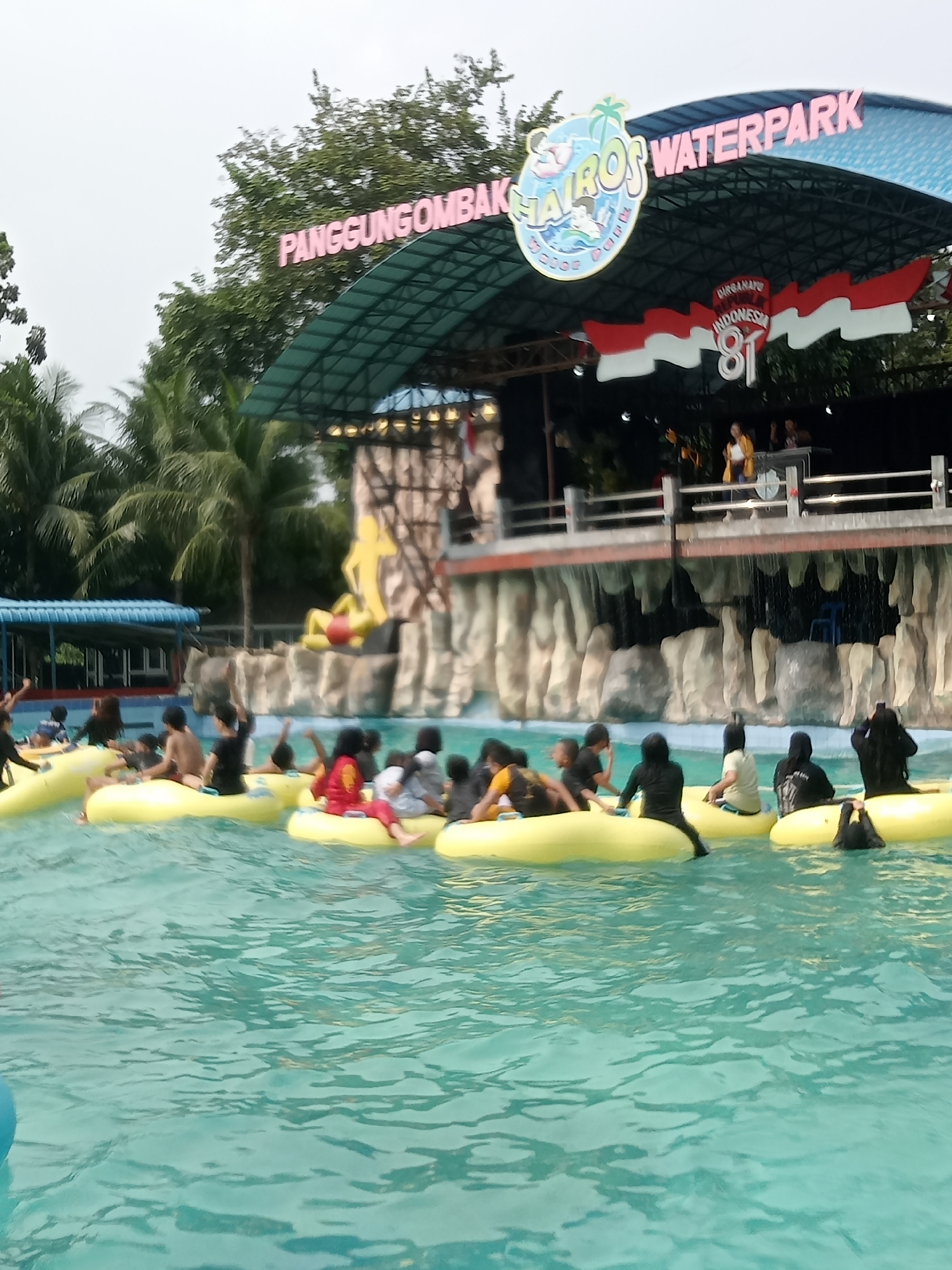 The image depicts a scene at a waterpark, identified by the signage "PANGGUNGGOMBAK AIROS WATERPARK" and the Indonesian flag. The waterpark appears to be located in the city of Baru, Indonesia. In the foreground, several people, mostly children and young adults, are enjoying themselves in a pool, floating on yellow inner tubes. They are wearing swimwear and some are raising their hands, suggesting a lively atmosphere. The water is a clear turquoise color. In the background, there is a structure with a curved roof supported by beams. This structure has a stage-like area with people performing. Further back, there are palm trees and lush greenery, indicating a tropical or subtropical environment. The overall impression is a recreational area with people engaged in water-based activities under an overcast sky.
