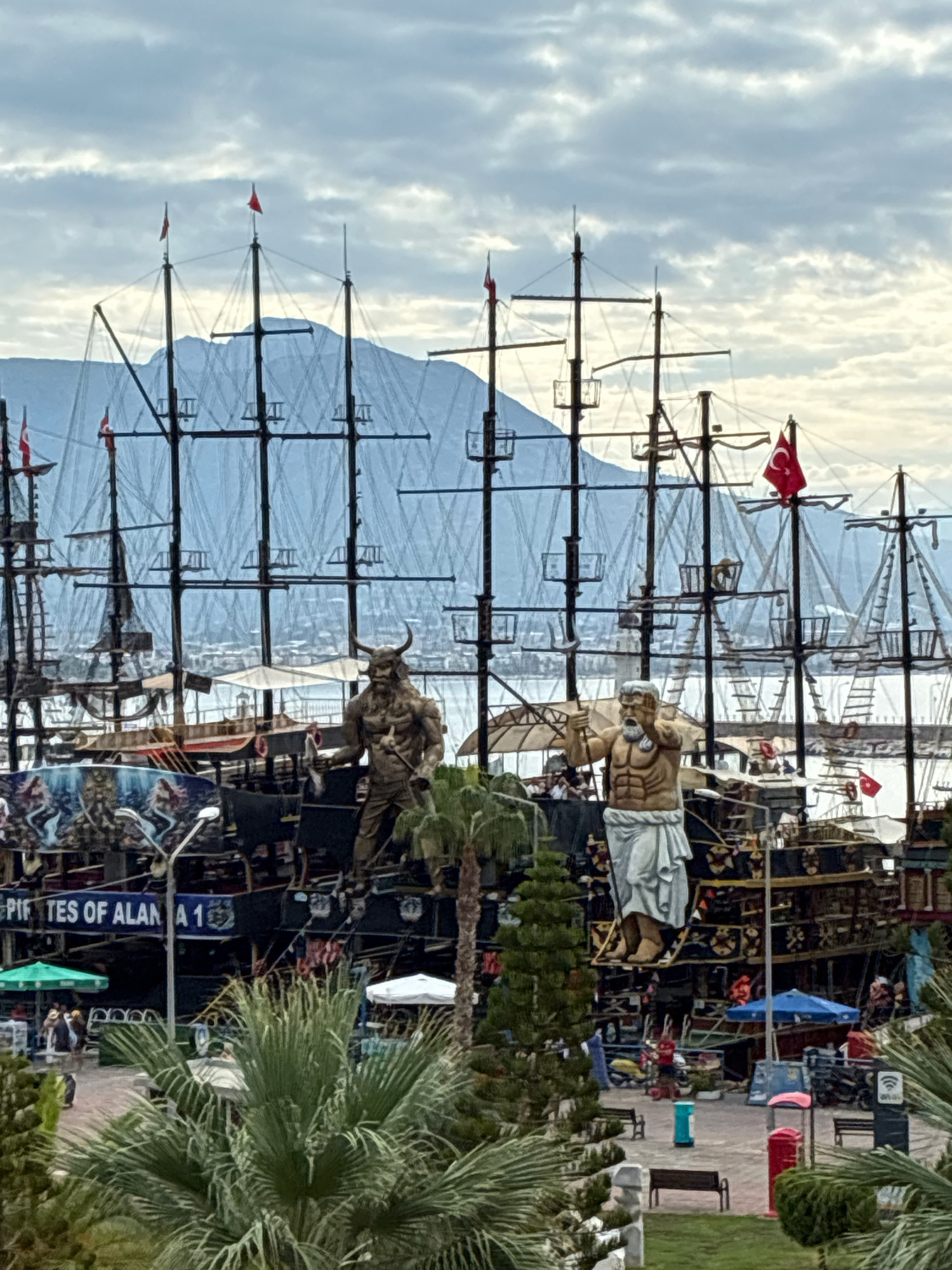 The image depicts a harbor scene in Alanya, Turkey, focusing on a fleet of elaborately decorated ships that appear to be themed as pirate vessels. In the foreground, lush green palm trees and other foliage obscure parts of the scene.
Dominating the mid-ground are two large, statuesque figures mounted on the decks of the ships. The figure on the left is a muscular, horned creature with a beard, resembling a minotaur. The figure on the right is a powerful, bearded man with an athletic build, wearing a white toga-like garment, and holding a trident. Both statues are rendered in a bronze or metallic finish.
The ships themselves are multi-masted with dark hulls, adorned with intricate designs and numerous small flags, including several red Turkish flags. Banners with text, one reading "PIF TES OF ALAN 1," are visible. Smaller boats and umbrellas are also present on the decks and in the surrounding area.
In the background, a misty mountain range rises behind the harbor, partially obscured by the ships' rigging and a cloudy sky. The water is calm, reflecting the structures. People are visible in the distance on the decks of the ships and on the shore, some appearing to be staff or tourists. The overall impression is a tourist attraction or entertainment venue designed to evoke a sense of adventure and history.