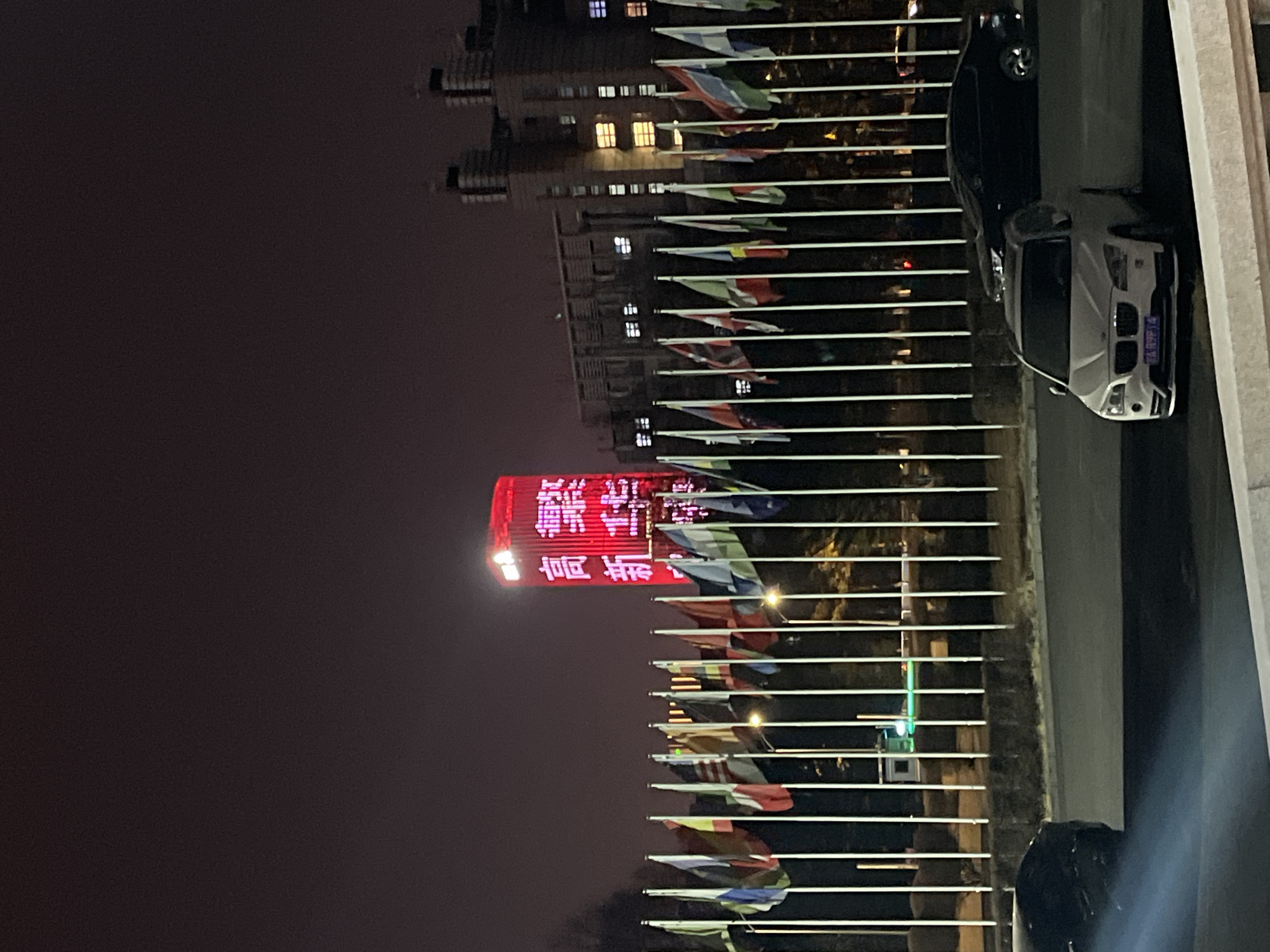 The image depicts a nighttime scene in Changchun City, China. A brightly lit red tower, displaying Chinese characters vertically, dominates the left side of the frame. Rows of international flags on poles extend horizontally from the right edge towards the tower. Two cars, a dark sedan and a light-colored SUV, are parked near a vertical concrete wall. The environment suggests an urban setting at night.
