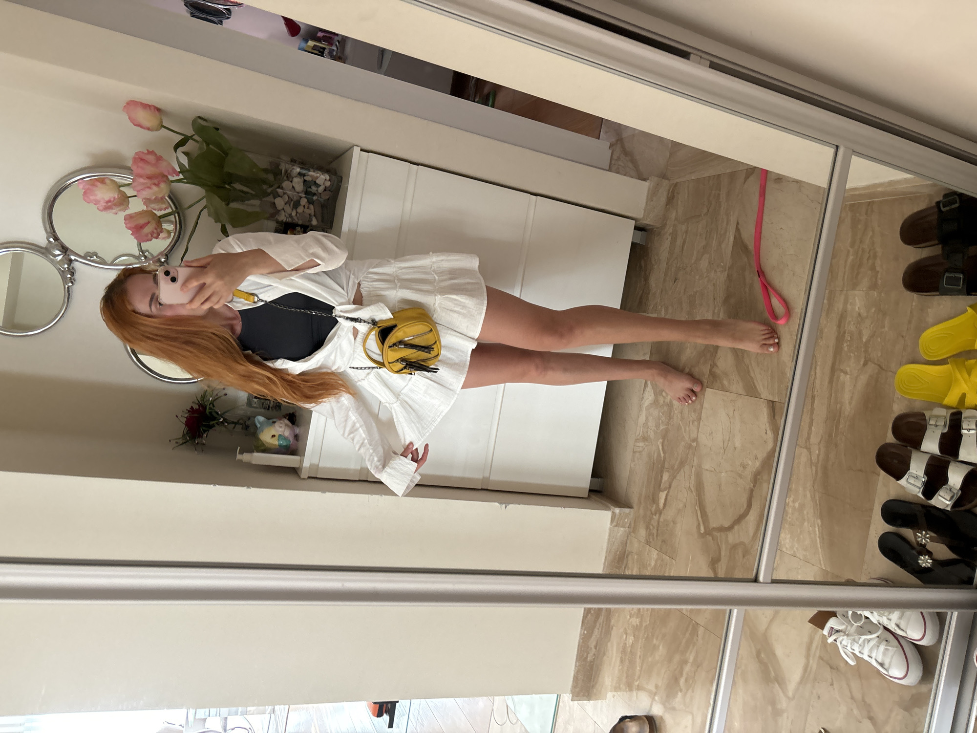 A person with long, auburn hair is taking a mirror selfie in what appears to be a bedroom or dressing area. They are wearing a white top with ruffled sleeves and a white skirt. A small yellow purse with a chain strap is slung across their body. The background shows a white dresser or closet, with decorative pink tulips in a vase on top. In the reflection of the mirror, several pairs of sandals and sneakers are visible on the floor. The lighting suggests it is daytime. The overall impression is casual and domestic.