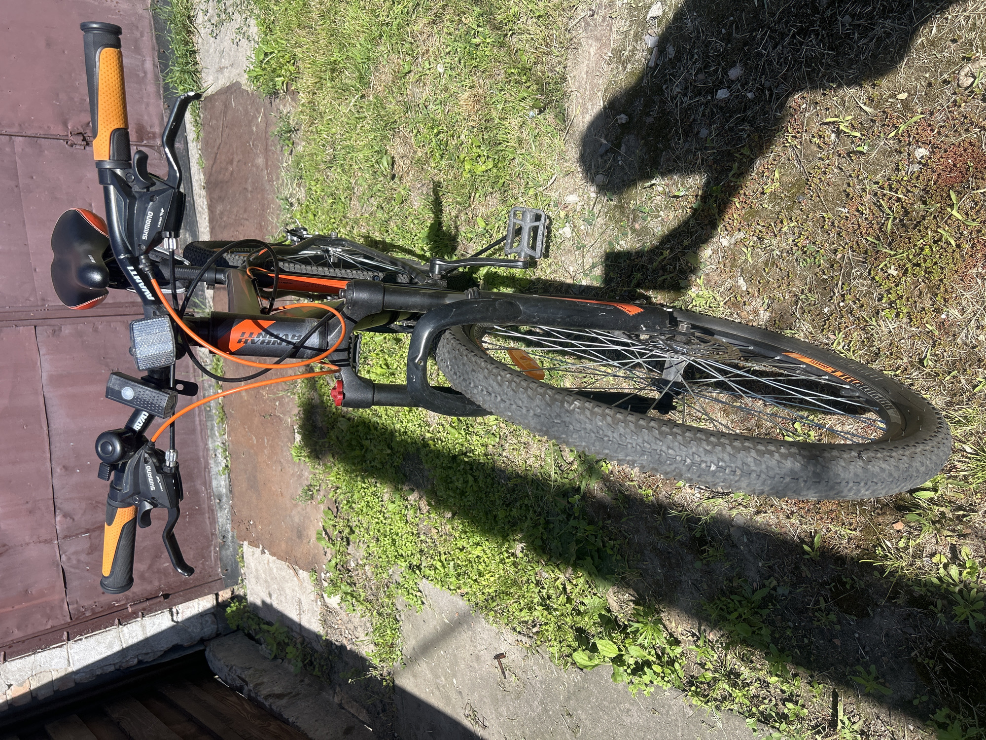 An orange and black Avanti bicycle is parked outdoors on a sunny day, casting a long shadow on the ground. The bicycle has orange handlebar grips, a black seat with orange trim, and a bell. A Shimano gear shifter is visible on the handlebars. The bicycle is parked on a patch of grass and dirt next to a concrete surface. In the background, there is a brown, corrugated metal door. The scene appears to be in a residential or back alley area. The location is Bila Tserkva, Ukraine.