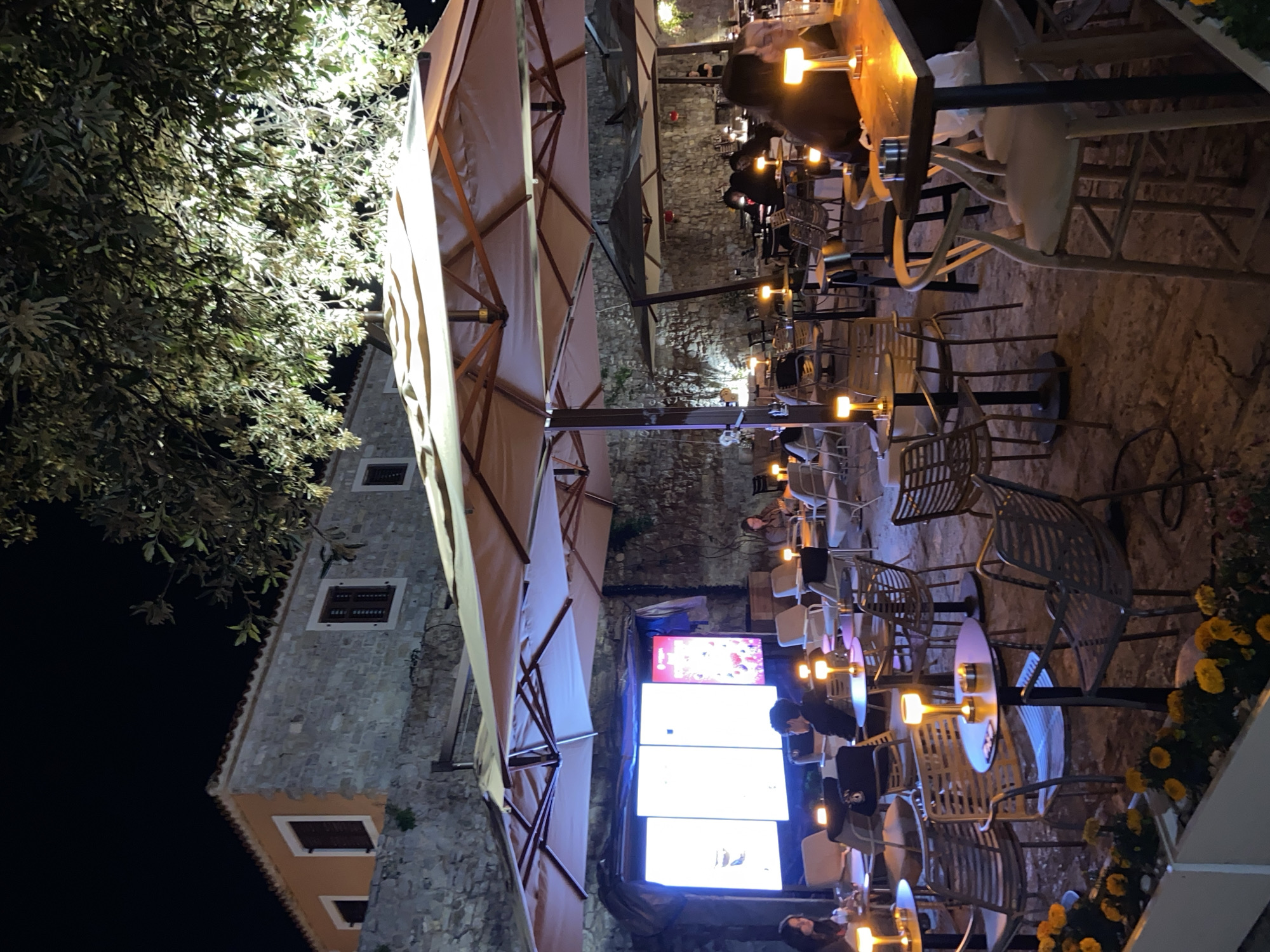 This image shows an outdoor cafe at night in Budva, Montenegro. The cafe is situated next to a stone building with multiple windows and is covered by large umbrellas. People are seated at tables, lit by small lamps. The cafe is decorated with yellow flowers. A large digital screen displays images or advertisements.