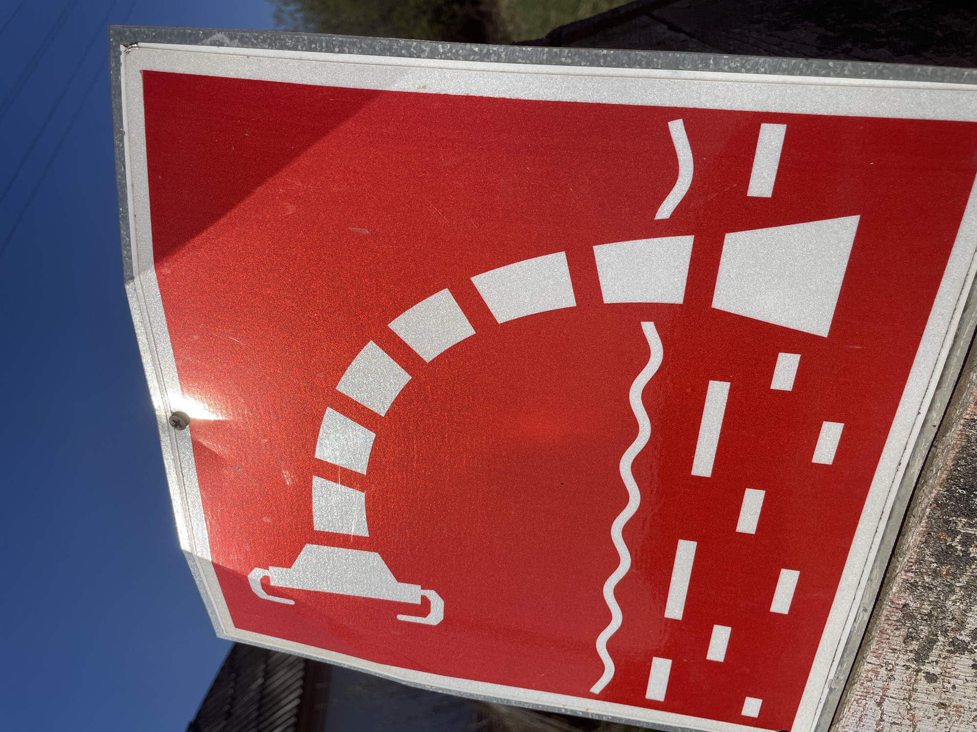 A close-up, angled shot depicts a red and white road sign. The sign, encased in a metal frame, is likely made of a reflective material given the strong sunlight reflection. The image on the sign features white symbols on a red background. The symbols depict a stylized bridge or viaduct, with a vehicle passing underneath. To the right of the bridge is a wavy vertical line, and below, a series of short, vertical lines resembling rain. The sign is mounted on a wooden surface. A portion of the clear blue sky is visible in the lower left corner of the image. The location is reported to be in Поедуги, Russia, though there are no inherent geographic markers in the image to confirm this.