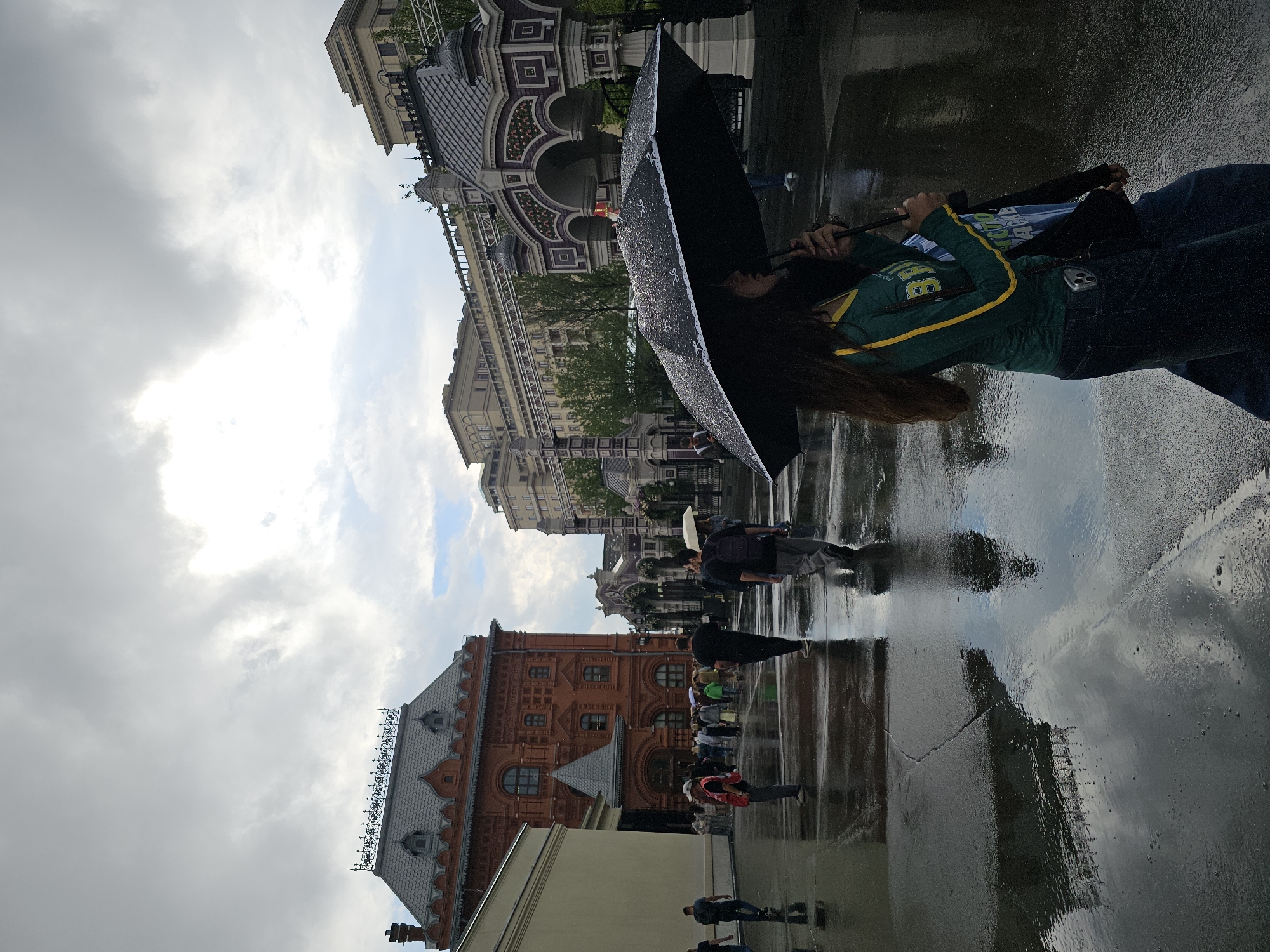 The image depicts a group of people standing on a wet, reflective surface, likely a street or plaza, under an overcast sky. The ground is heavily saturated, indicating recent rain. A person in the foreground is holding a large black umbrella open, sheltering themselves and possibly others. This individual is wearing a green long-sleeved shirt with yellow trim and a blue shirt underneath, the latter bearing some white text.
In the background, there are several historic buildings with distinctive architectural styles. One building, prominent on the right, is made of red brick with arched windows and decorative rooflines. To its left and further back, another building features ornate facades with arched entrances and patterned detailing. The overall architecture suggests a European city, and the context provided indicates Moscow, Russia.
The lighting is diffused, consistent with a cloudy or rainy day. The reflections on the wet ground create a mirrored image of the surroundings, adding depth to the scene. The mood appears to be subdued, likely due to the weather. Several other individuals are visible, some holding umbrellas, walking or standing in groups. The activity seems to be people navigating the area in rainy conditions, possibly as tourists or residents going about their day. The presence of multiple people suggests a public space.
