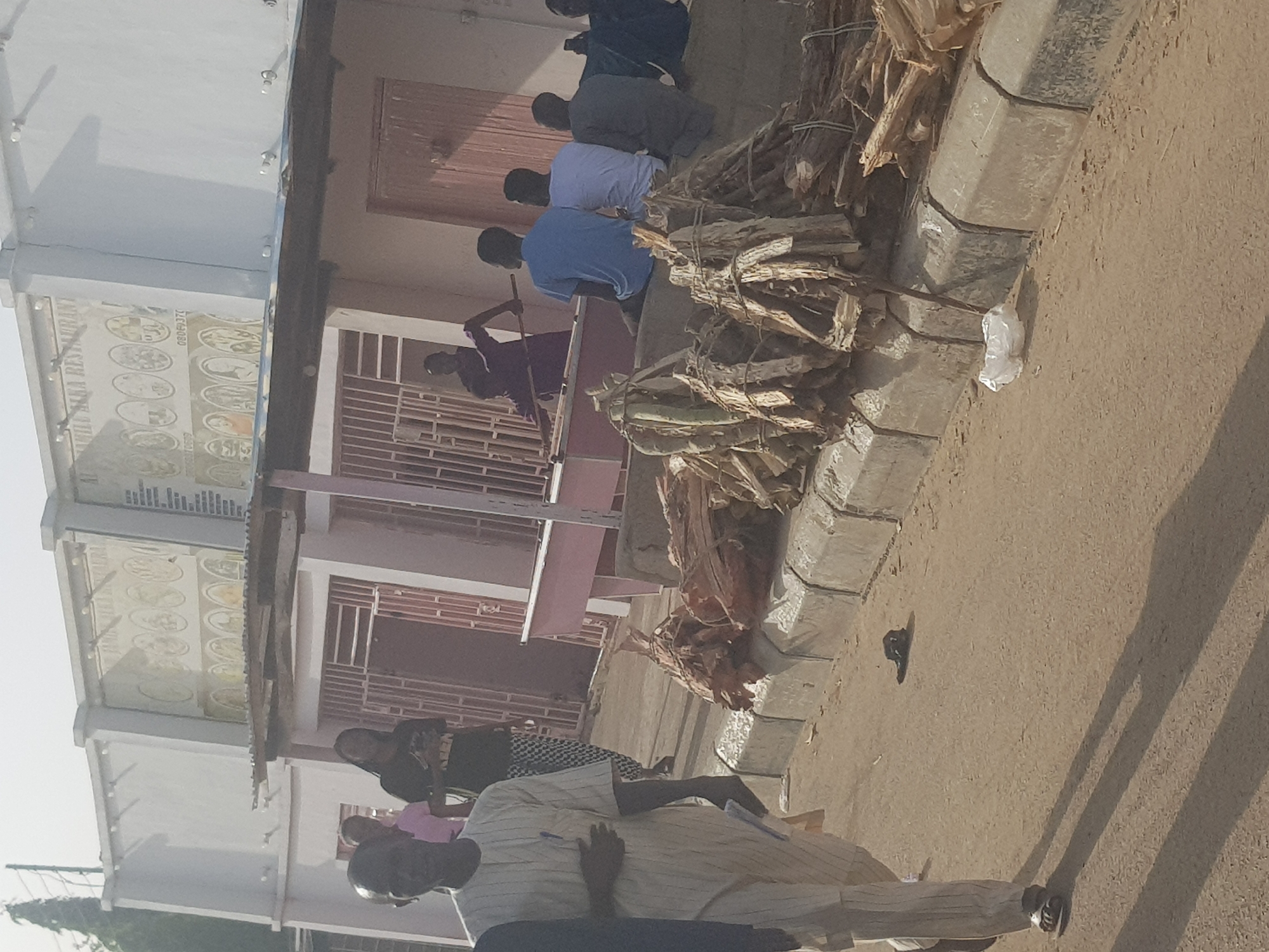 Location: Dabawa, Nigeria.
Scene: A row of people sitting outdoors next to bundles of tied sticks/wood. One person is standing, appearing to be working with a tool near the wood. The bundles are stacked along the edge of a sidewalk. A building with a shopfront sign is in the background. A person is walking on the foreground. The ground is dirt/sand.
