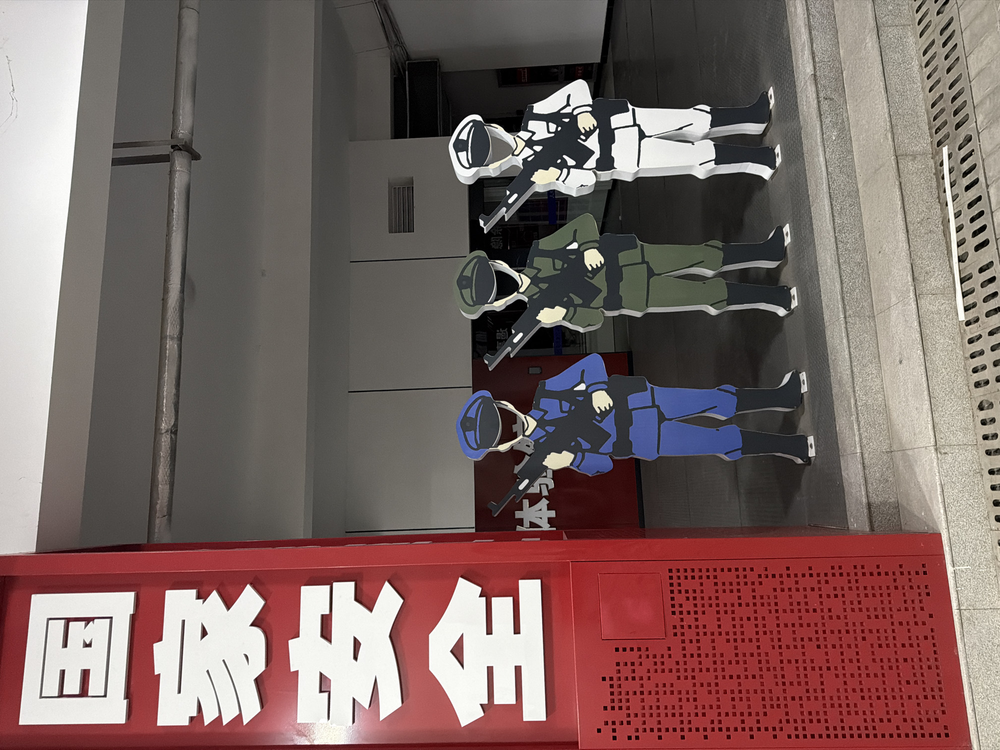The image captures an indoor or covered outdoor area in Jiangning District, China. A prominent red vertical sign on the left displays "国家安全" (National Security) in large white Chinese characters, with its lower section featuring a pattern of small square perforations. To the right of this sign, three two-dimensional, cartoon-style cutout figures are positioned in a line on a light-colored tiled step. Each figure, depicted with a blank black facial area and wearing a cap, gloves, and boots, holds a black firearm horizontally across its chest, pointed rightward. From left to right, the figures are uniformly colored blue, olive green, and white. The background consists of a light-toned wall or interior space, revealing a horizontal pipe near the ceiling and ventilation grates. The foreground shows a metal drainage grate on the ground level, with a thin white strip of material lying horizontally on it. The scene is evenly illuminated.