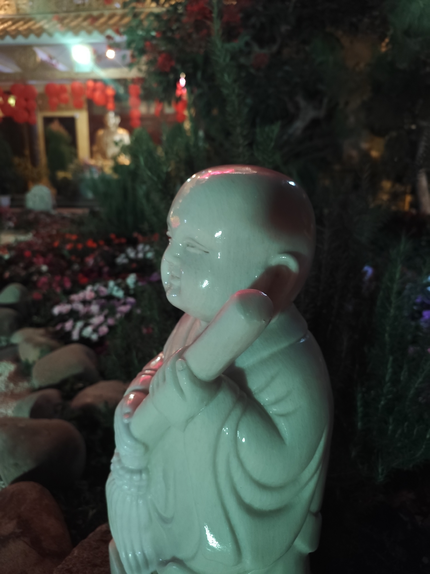 The image shows a small, light-colored statue of a young, bald figure, possibly a child monk, holding a staff over their shoulder. The statue is the central focus, positioned close to the camera. In the background, blurred and out of focus, are elements suggesting a garden or temple setting. These include flowers, rocks, greenery, and what appear to be red lanterns and a structure possibly associated with a temple.
The scene is likely at night, given the soft, diffused lighting and the illuminated lanterns. There is no immediately discernible evidence that the location is Da Lat, Vietnam, but the elements in the background (temple architecture, garden layout, lantern style) could align with gardens or temples found in that city.