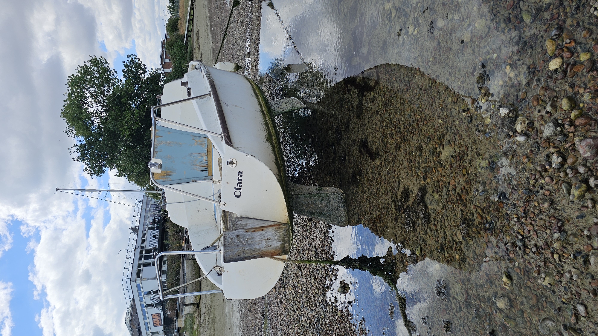 A white sailboat named "Clara" is beached on a rocky shore at low tide. The boat, tilted to its starboard side, rests on a concrete keel block. A wooden plank is attached to the stern of the boat. The background features a cloudy sky, a modern building with "ADUR" signage, and green trees. The foreground is dominated by the rocky beach and shallow water reflecting the sky and the boat.