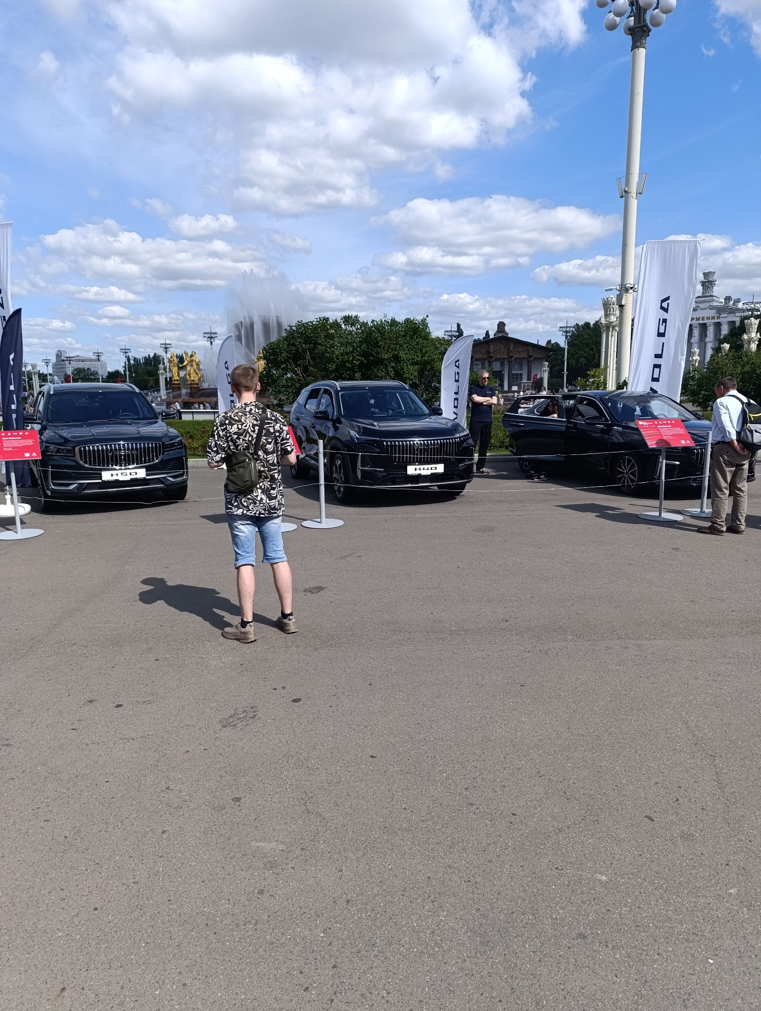 The image captures an outdoor exhibition or car show, likely at VDNKh in Moscow, Russia, as suggested by the architecture and the presence of fountains in the background. The scene is bathed in daylight under a sky with scattered clouds. In the foreground, an asphalt surface is visible, with a young man in a patterned shirt and denim shorts standing center frame, looking towards the right. He is wearing a cross-body bag. To his left, a dark-colored SUV, identified by the "K50" emblem on its grille, is parked. To the right of the young man, another dark-colored car, a sedan with its doors open, is displayed. This car has a red informational display in front of it. Several tall white banners with the "VOLGA" logo are positioned around the exhibition area. In the middle ground, more of the VDNKh park's distinctive architecture, including large fountains and golden statues, can be seen. A few other individuals are present, including two men near the Volga banners and another person partially visible inside the open-doored sedan. The overall impression is a public event showcasing vehicles in a well-known Moscow location.