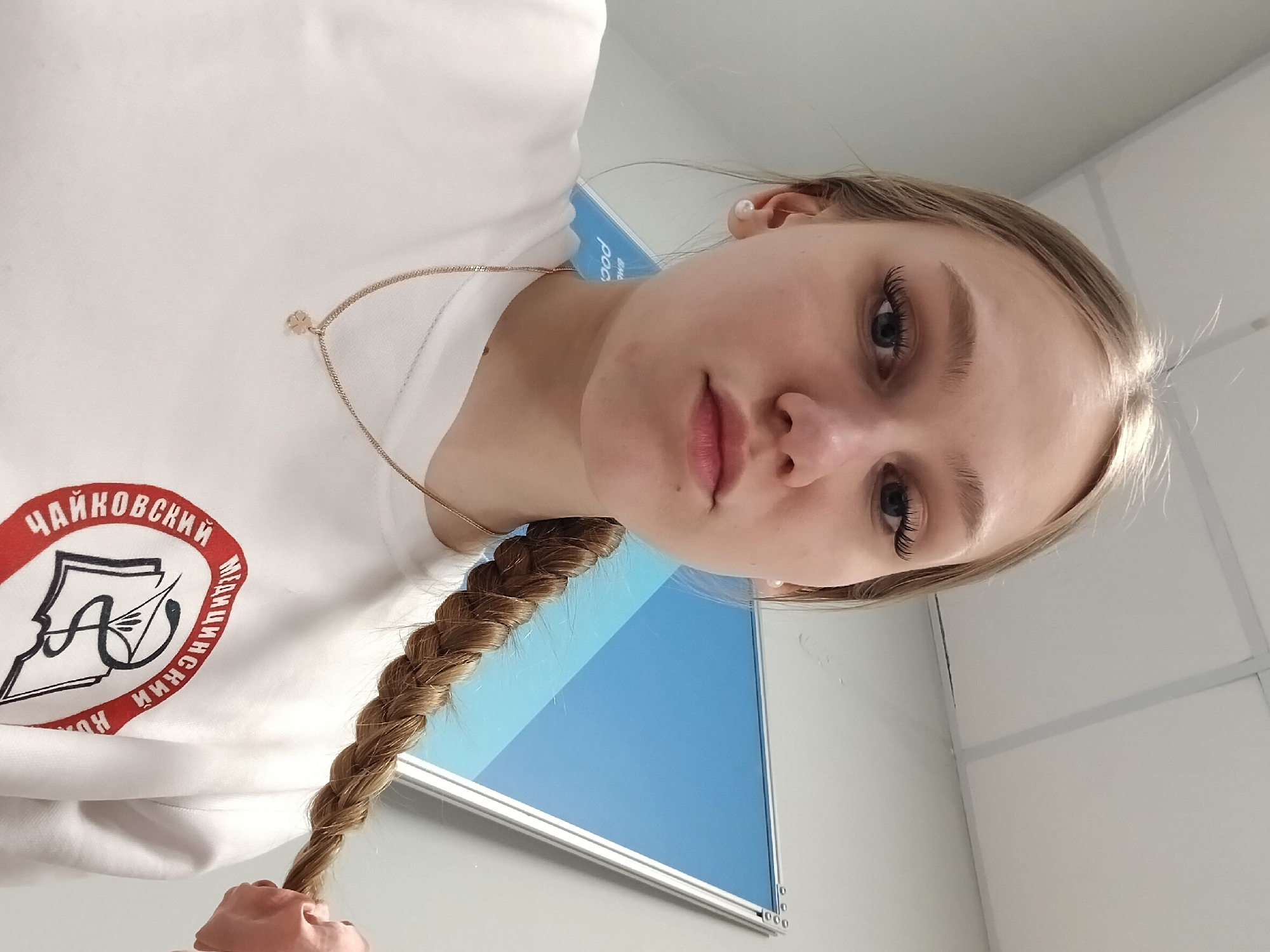 A fair-skinned young woman with long blonde hair styled in a braid is pictured indoors, likely within a building in Perm, Russia. She's wearing a white sweatshirt with a red circular emblem featuring text in Russian, identifying the city of Chaikovsky, along with imagery that appears to be related to music and possibly a school. She also wears a delicate gold chain with a small charm. A blue panel or surface with a metallic frame is visible behind her, possibly a partition or informational display. The background suggests an office or institutional environment with white walls and ceiling tiles.