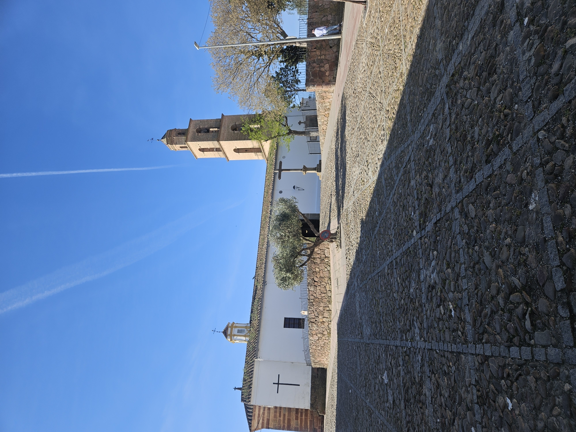 The image depicts a view of the Iglesia de Nuestra Señora de la Asunción, a church located in the town of Pedro Abad, Spain. The church features a tall, multi-tiered brick bell tower, which is the dominant vertical element in the scene. The main body of the church is white with brick accents, and a large black cross is painted on one of its walls. In front of the church is a stone cross monument. A cobblestone street occupies the foreground, with a shadow cast across a portion of it. The sky is clear blue, with contrails visible overhead. A mature tree is positioned between the street and the church. Along the roadside, a person is standing next to a light pole on the street, near a wall.